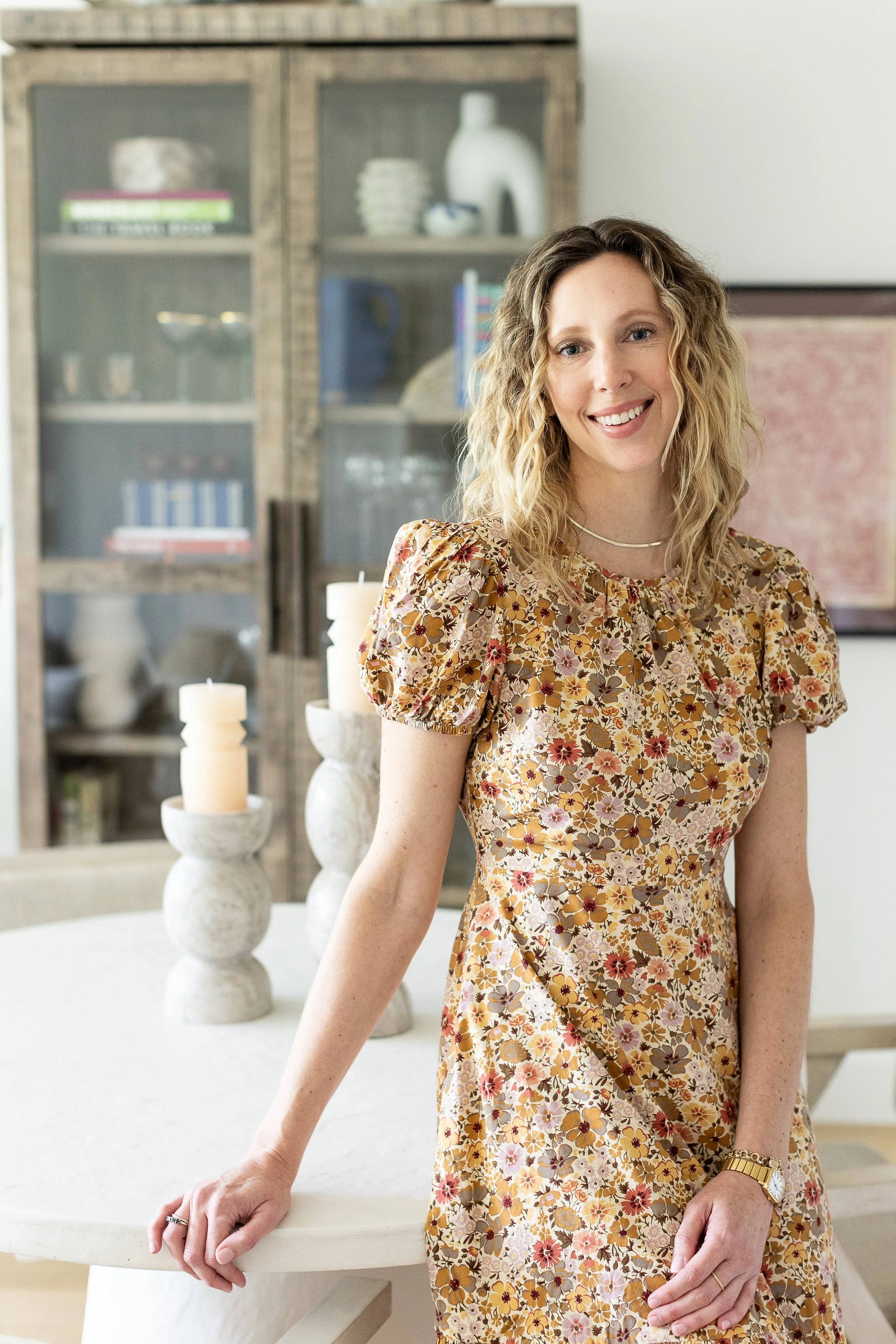 A smiling woman with wavy blonde hair wearing a floral dress, standing indoors next to a white table with candles in the background.