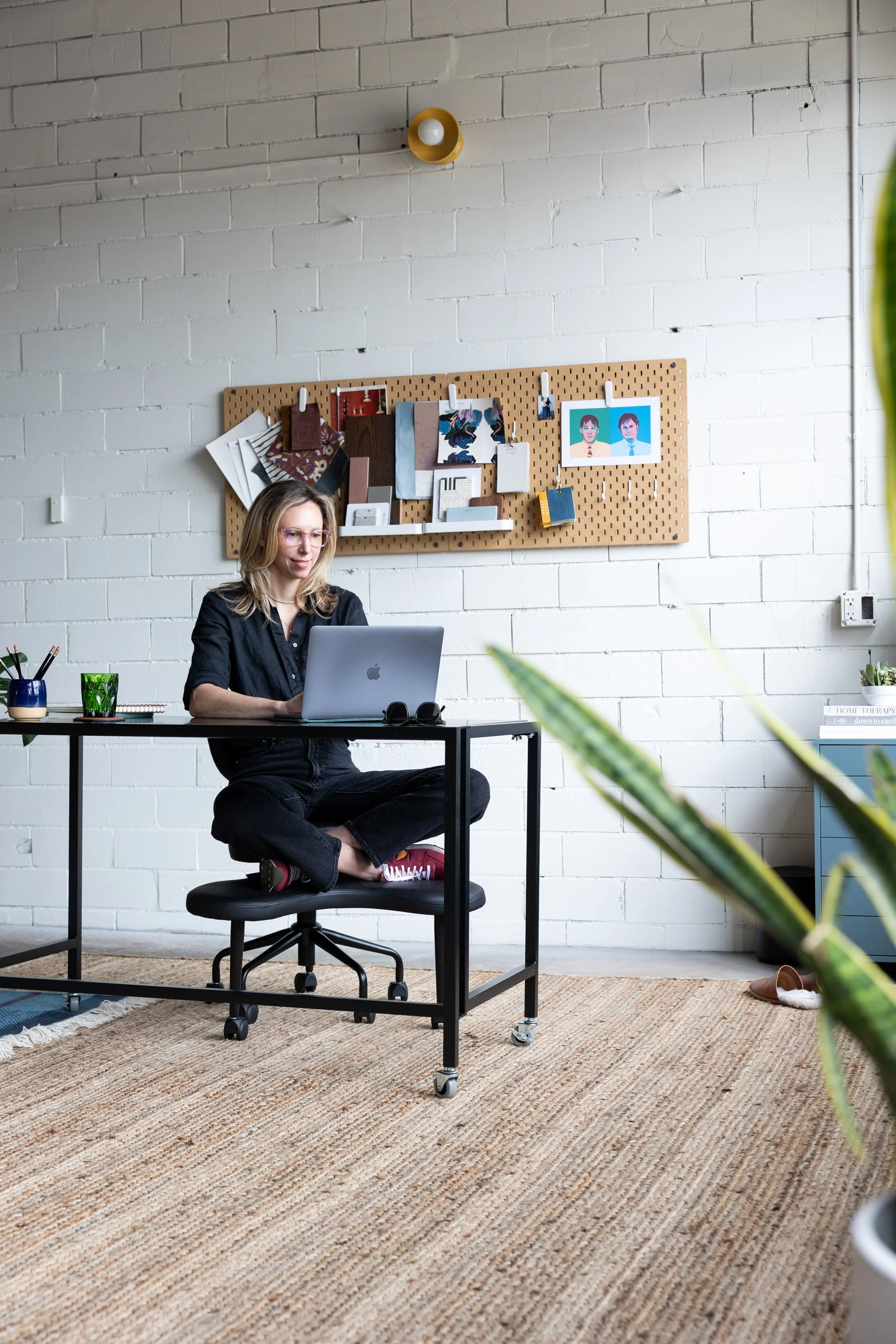 A woman with blonde hair and pink glasses sitting cross-legged on a rolling office chair at a desk, working on her laptop. The room has a white brick wall with a pegboard displaying various papers and art, a beige carpet, some house plants, and a small side table with books and decorations.