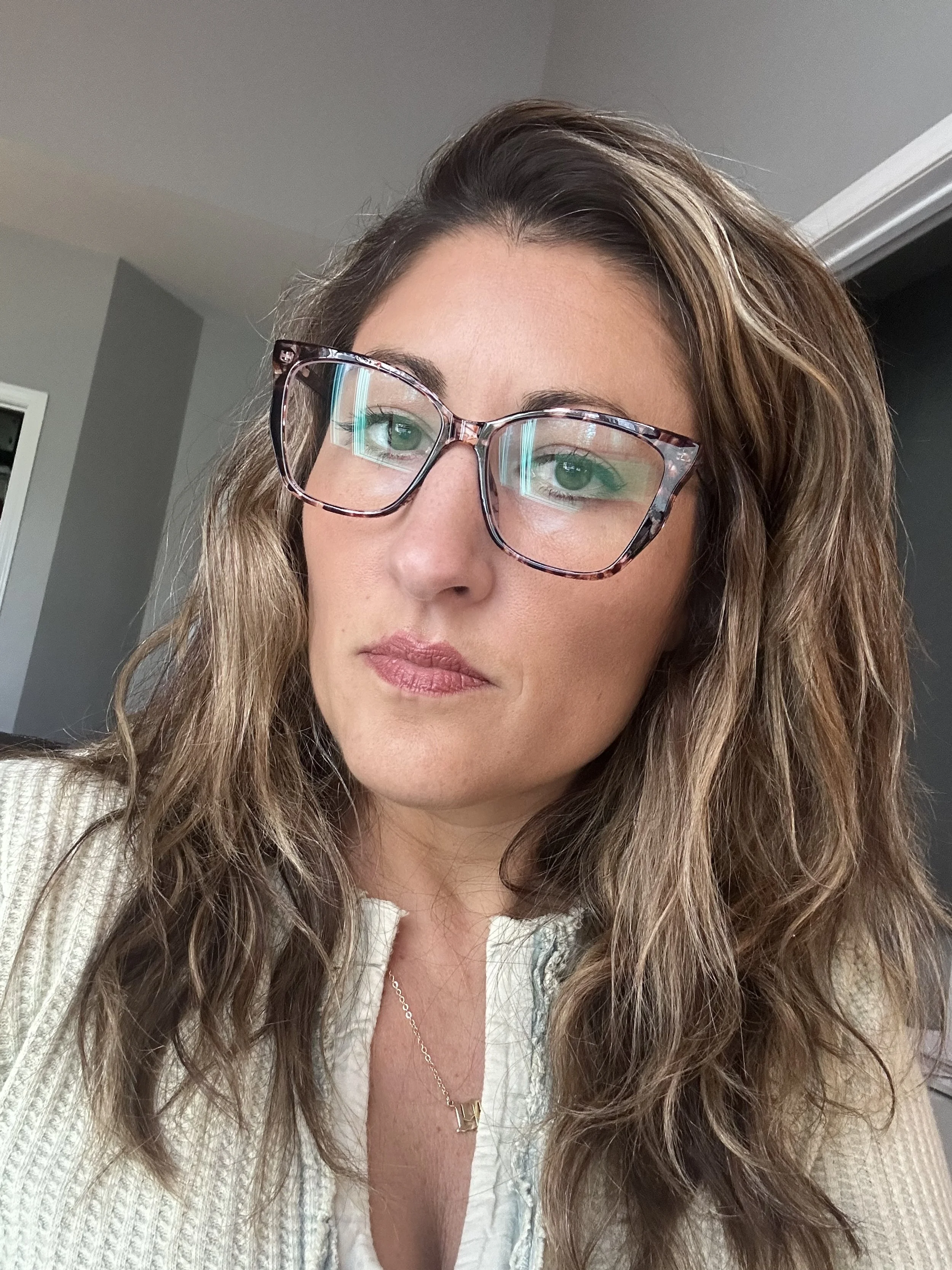 A woman with wavy brown hair wearing glasses with a tortoiseshell pattern, a light-colored sweater, and a gold necklace, taking a selfie indoors.