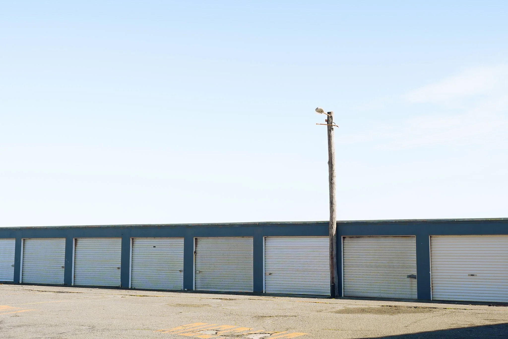 A row of closed gray garage doors in a commercial building with a single utility pole in front, and a clear sky above.