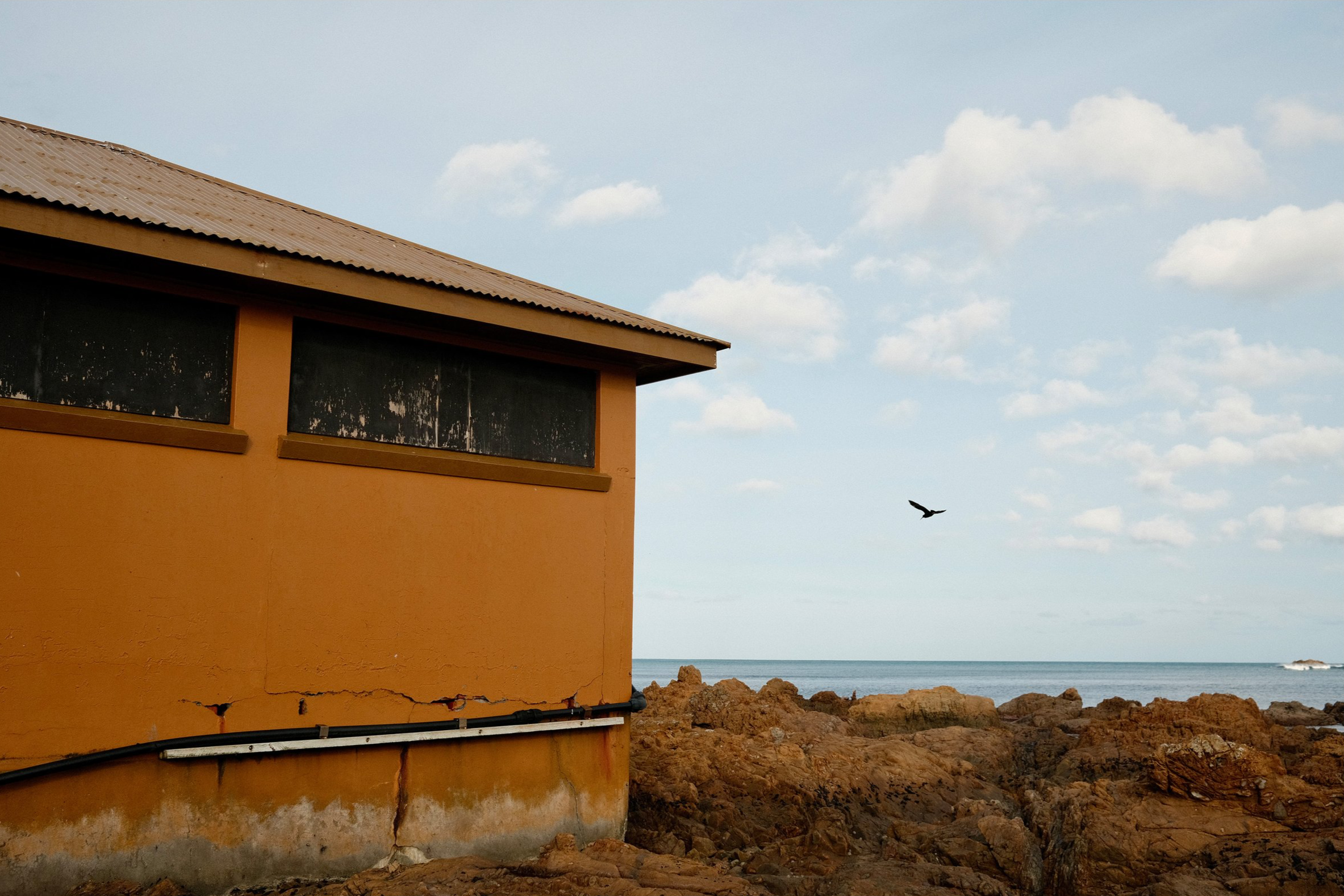 A building with a rusty metal roof, tan and black exterior walls, and a crack running down the lower part of the wall, situated by a rocky shoreline with the ocean and cloudy sky in the background.