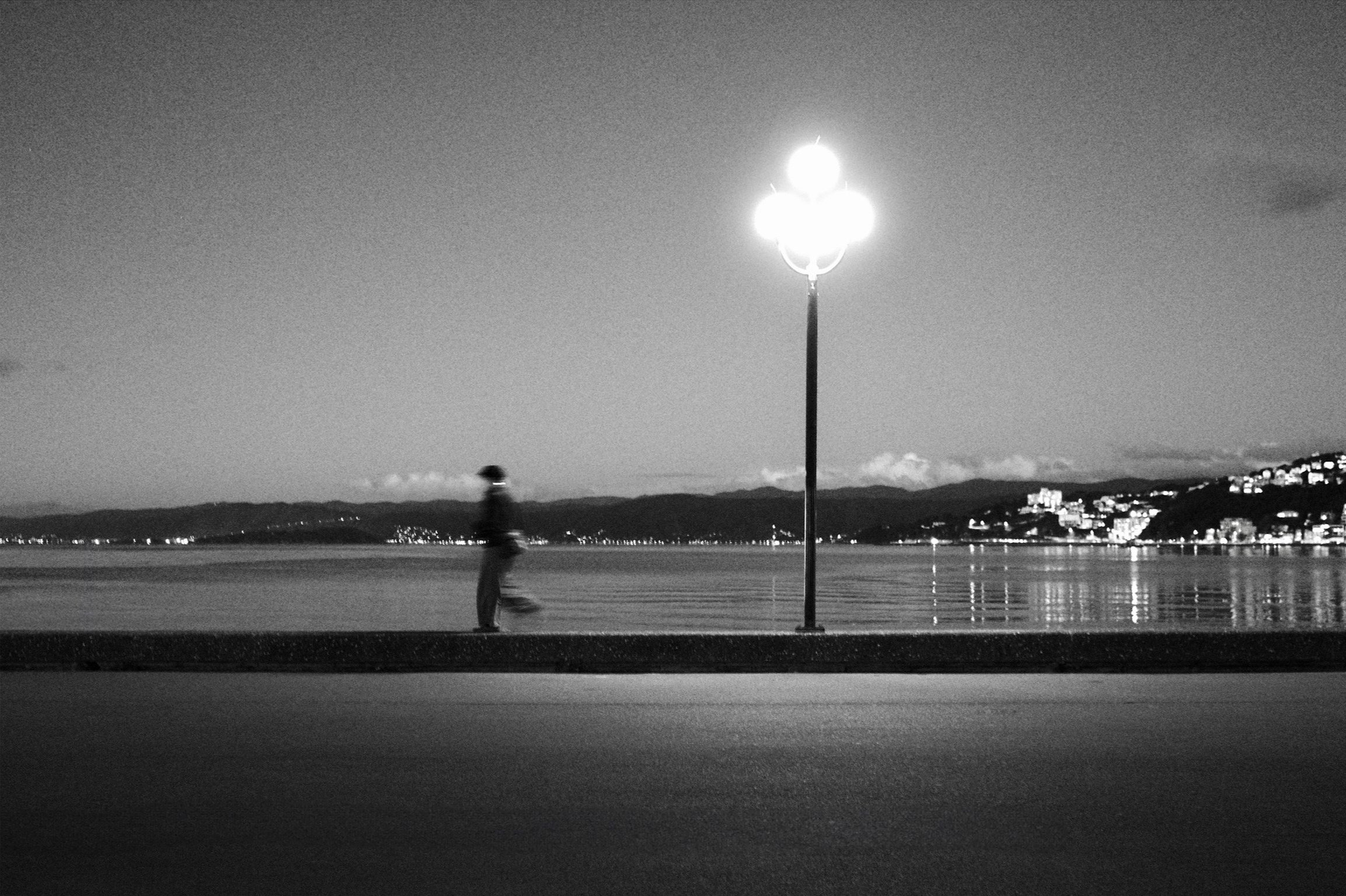 A black and white photo of a street lamp by a waterfront with a person walking past it. In the background, there are city buildings and mountains under a cloudy sky.