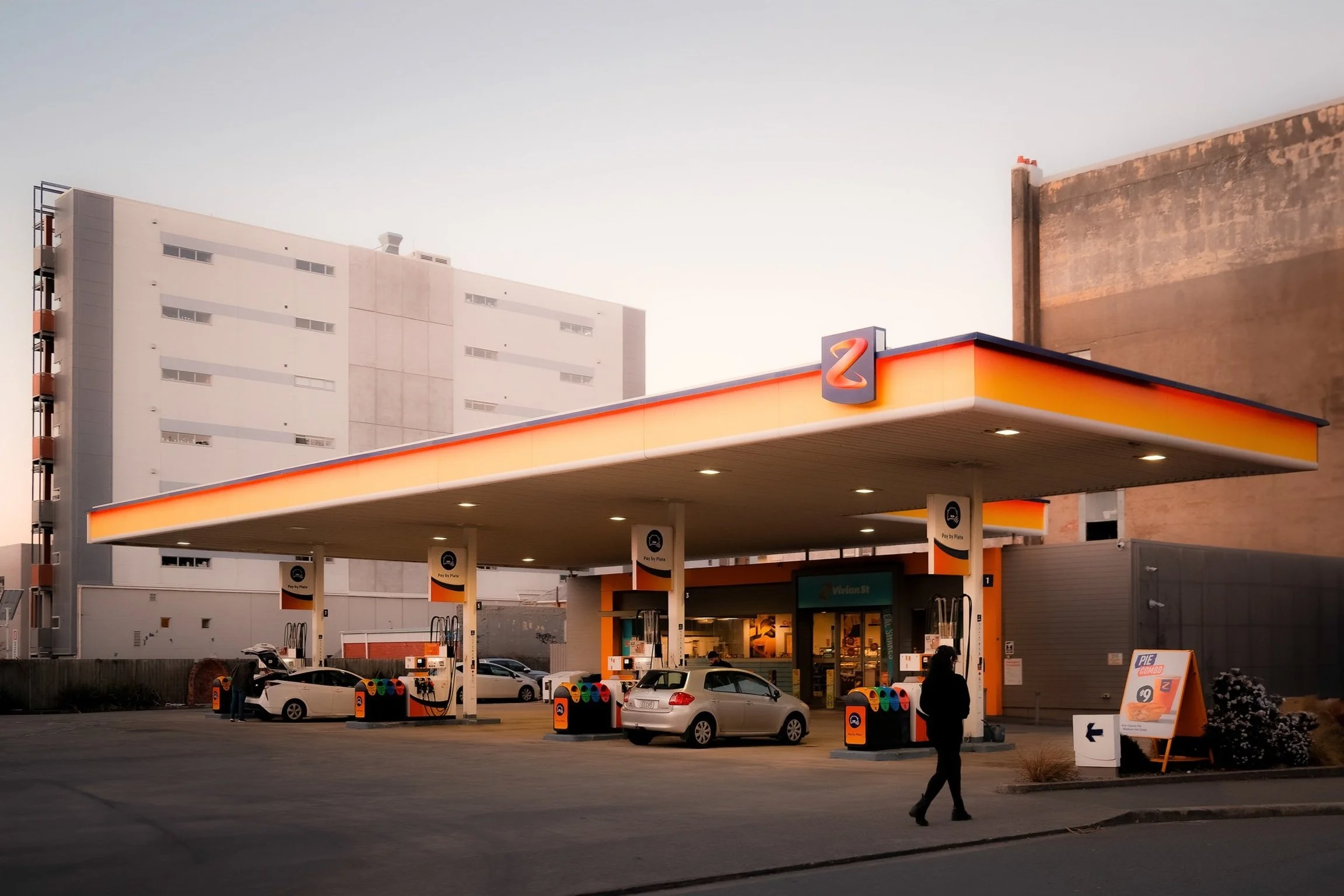 A Valero gas station with multiple fuel pumps and cars parked at the pumps. The station has a bright orange and yellow canopy with a Valero logo. There are tall buildings in the background and a person walking near the gas station.