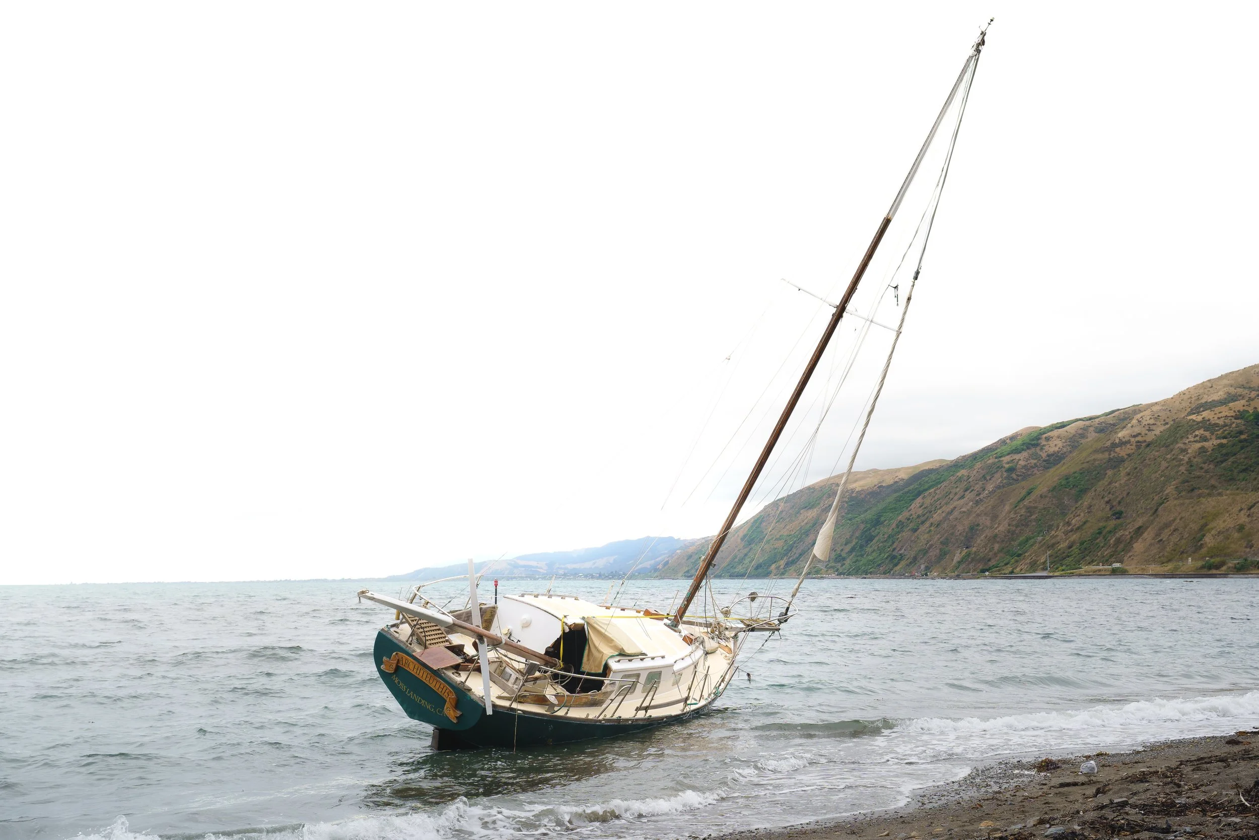A sailboat tilted on the water near a rocky shoreline with green mountains and cloudy sky in the background.