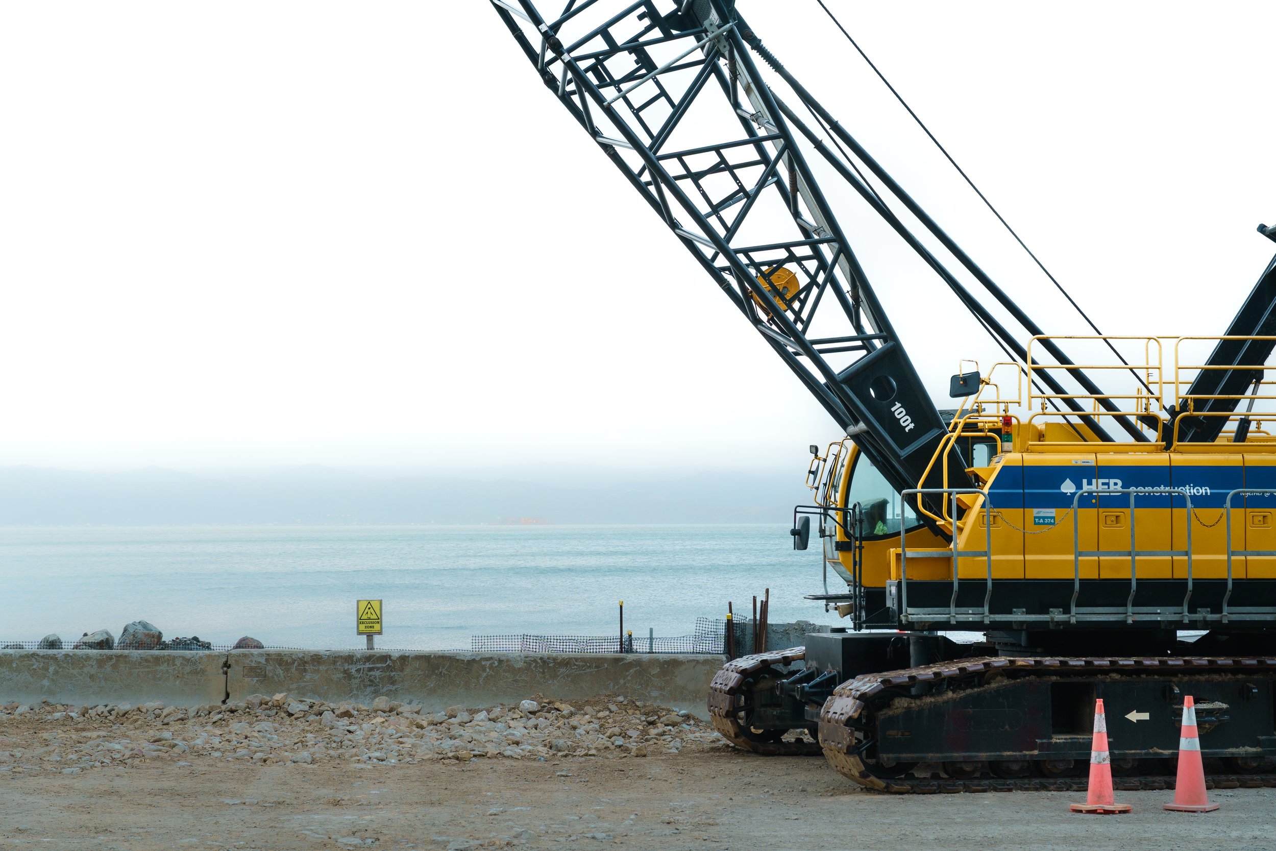 Construction excavator with tracked wheels on rocky terrain beside a body of water, with overcast sky and safety cones in the foreground.