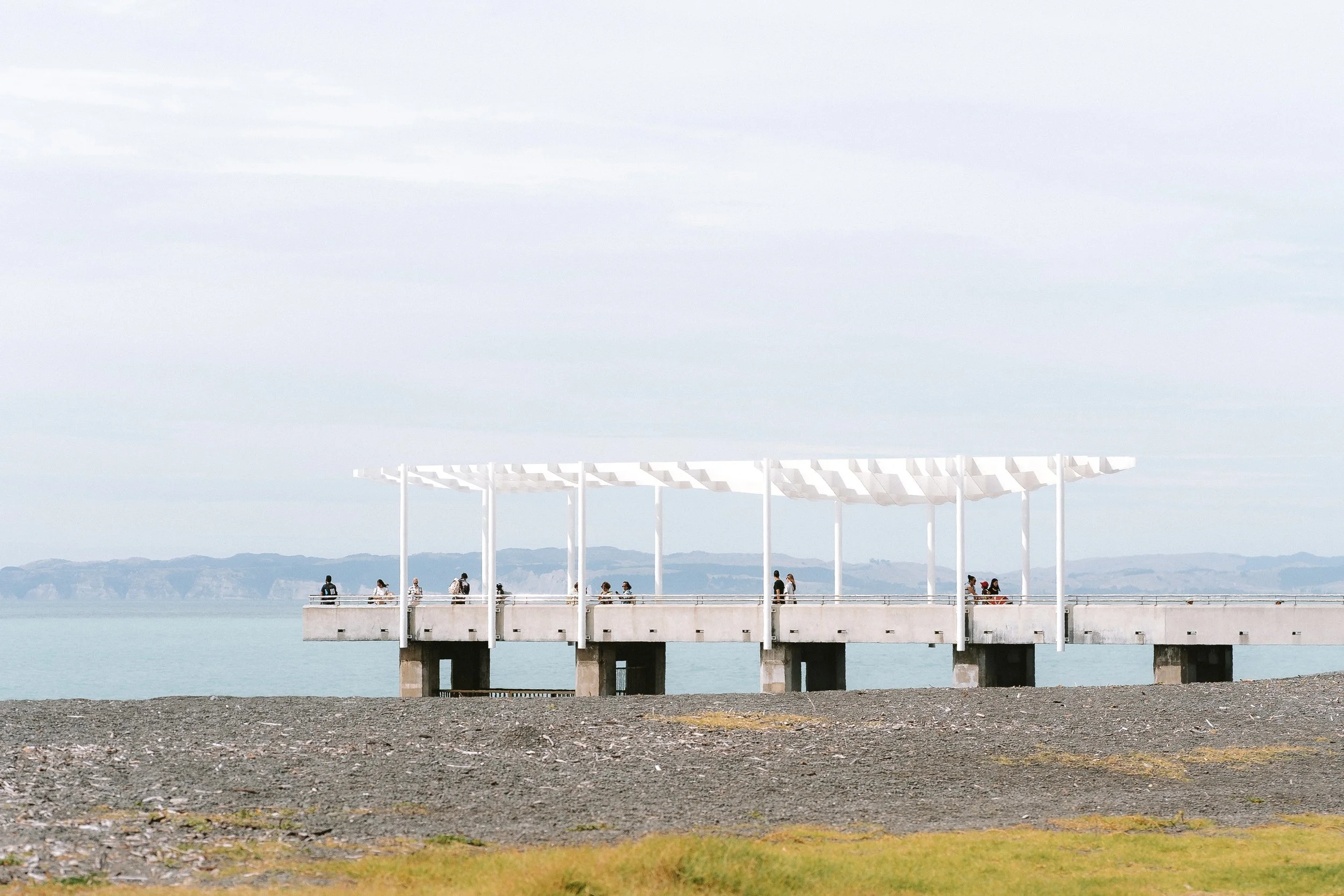 People walking on a pier with a white canopy structure over it, by a calm body of water under a cloudy sky, with distant mountains in the background.
