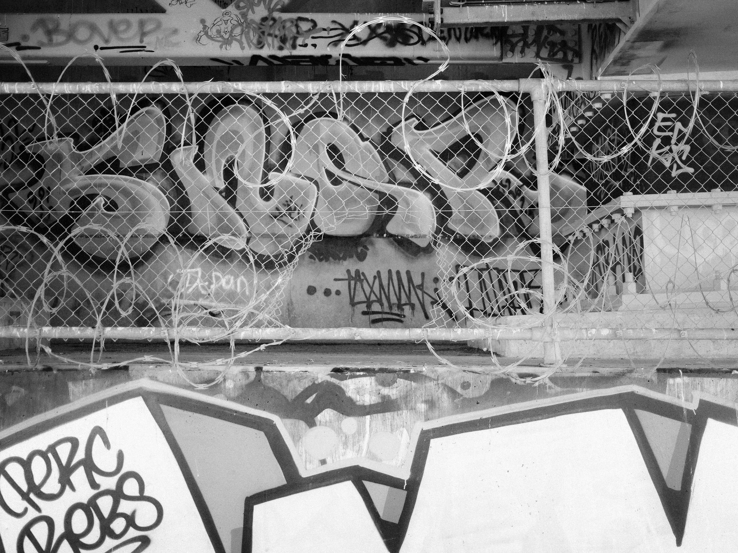 A fence with barbed wire on top, behind which are graffiti-covered walls and street art, including tags and cartoonish characters.