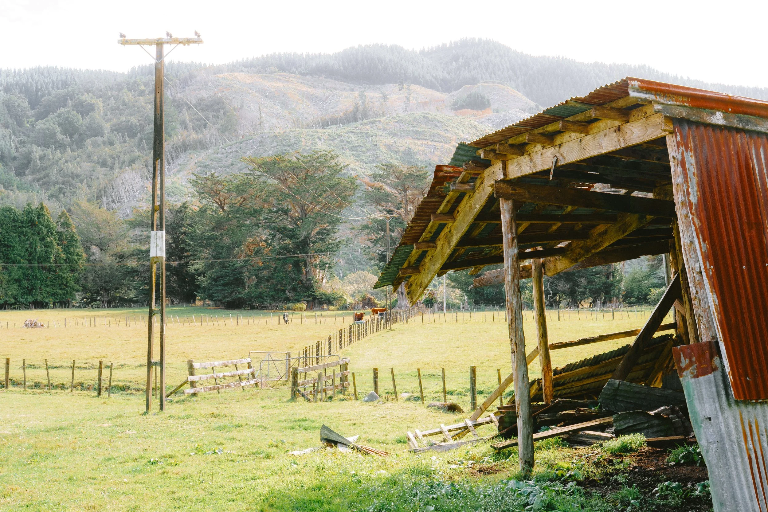 Rusty metal shed with a slanted roof in a rural farm area, green grass, wooden fencing, trees, and a mountain background.