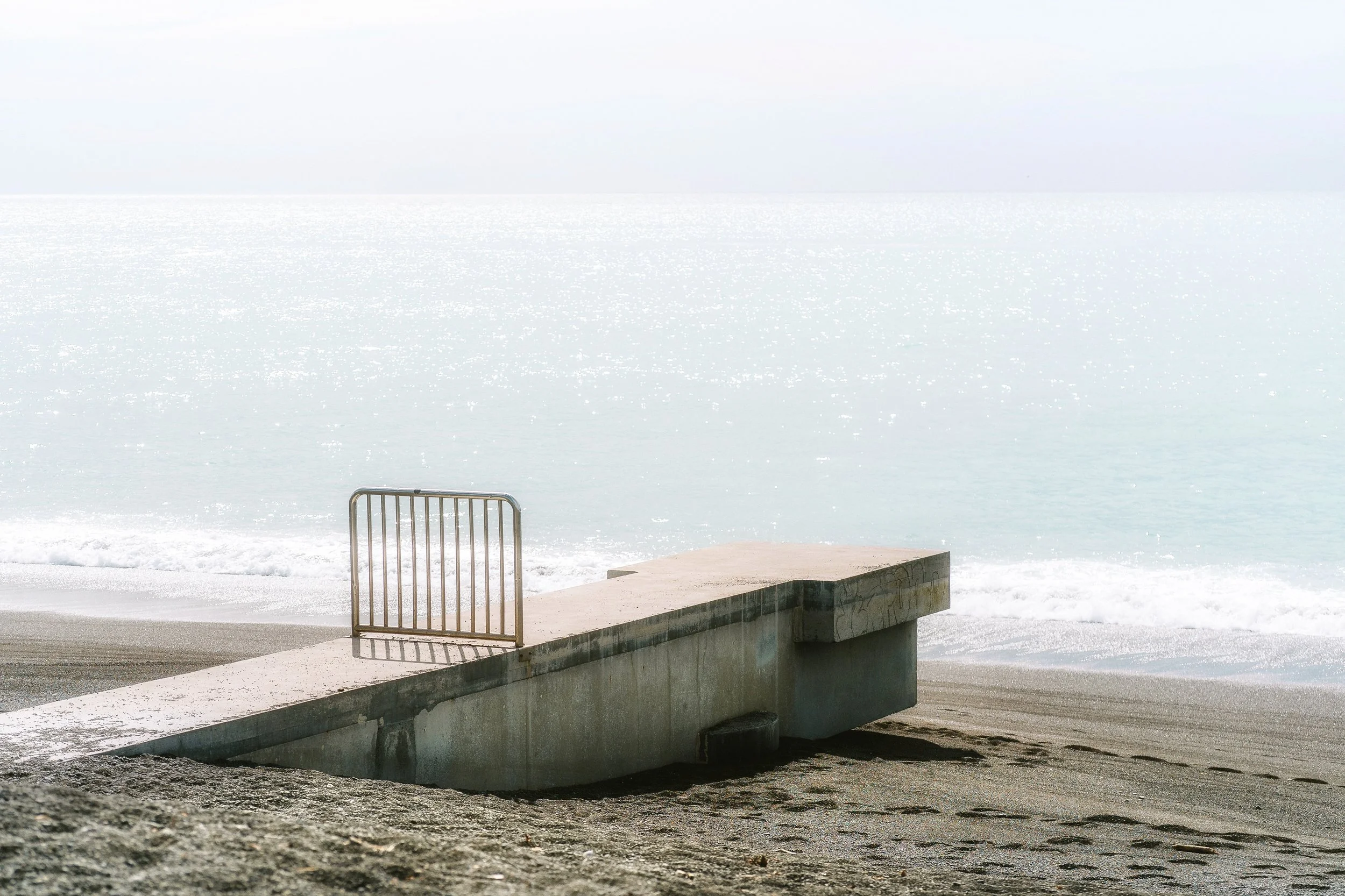 Concrete pier extending into a body of water with a metal railing, calm waters reflecting sunlight, and a light sky overhead.