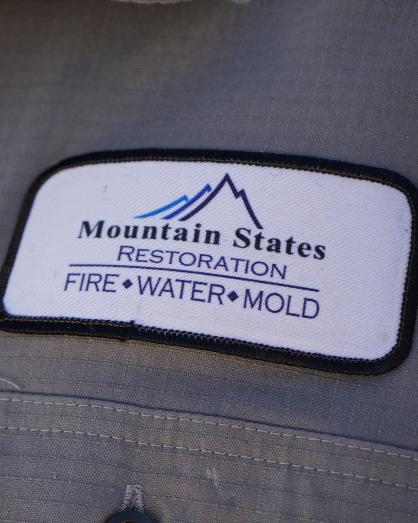 Close-up of a fabric patch with a mountain logo and the text 'Mountain States Restoration' and 'Fire Water Mold' on a gray fabric surface.