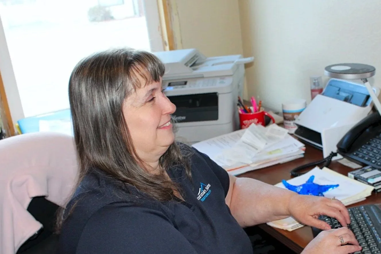 A woman sitting at a cluttered office desk typing on a keyboard, smiling, with a window, a printer, and various office supplies behind her.