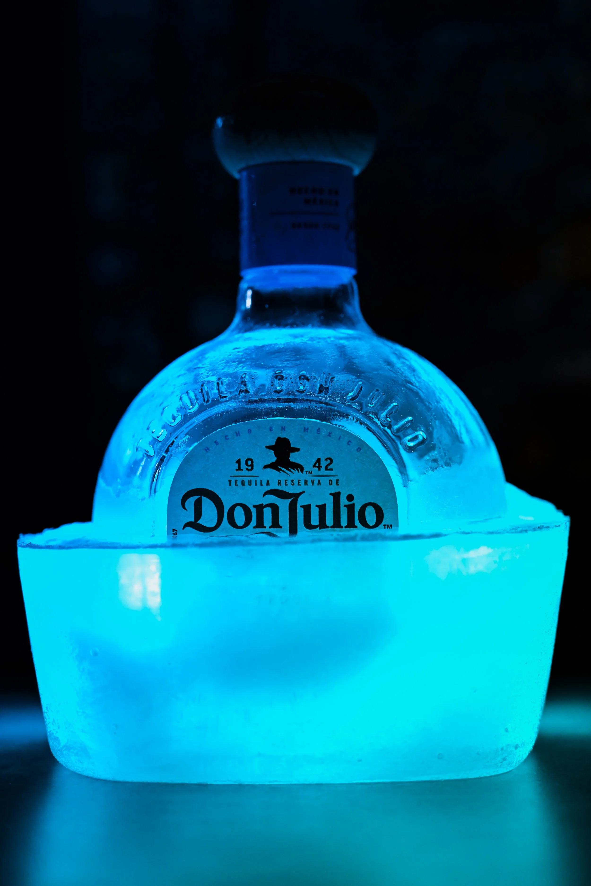 A clear glass bottle of Don Julio tequila, illuminated with blue light, sitting in a glowing ice bucket.