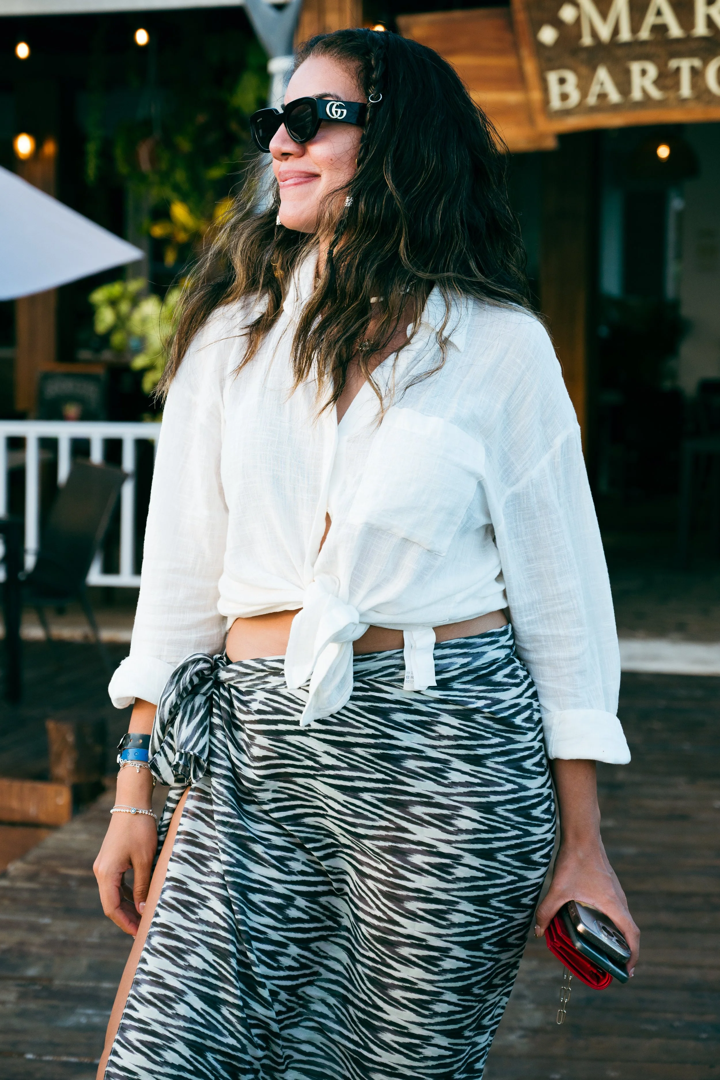 A woman with curly dark hair wearing large black sunglasses, a white tied-up shirt, and a zebra-print skirt, holding a phone and standing outdoors near a wooden building.
