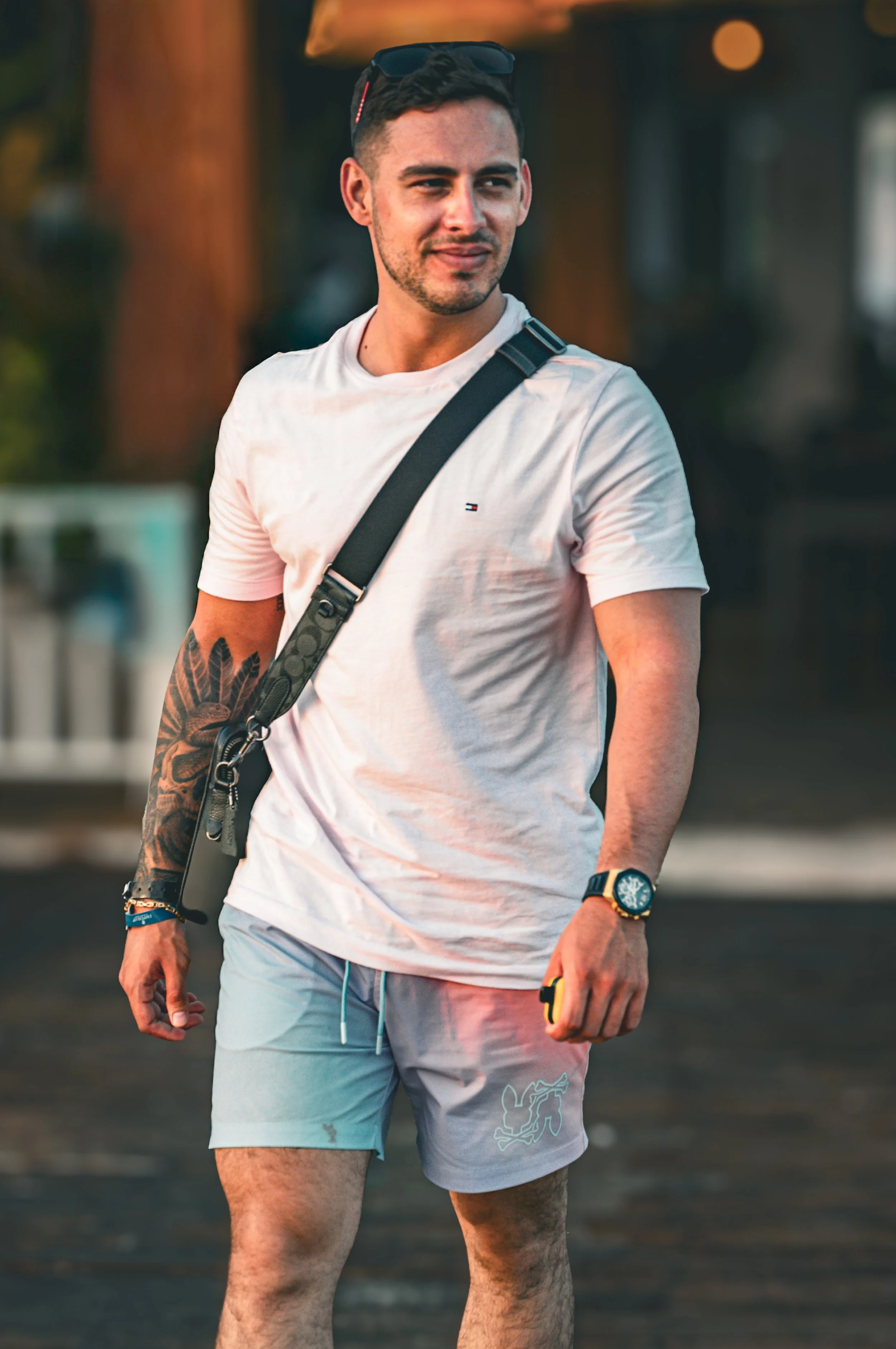 A young man with short dark hair, light skin, and tattoos on his left arm, standing outdoors. He's wearing a white t-shirt, light-colored shorts, a watch, and sunglasses on his head, carrying a black shoulder bag, and holding a small yellow item in his right hand.