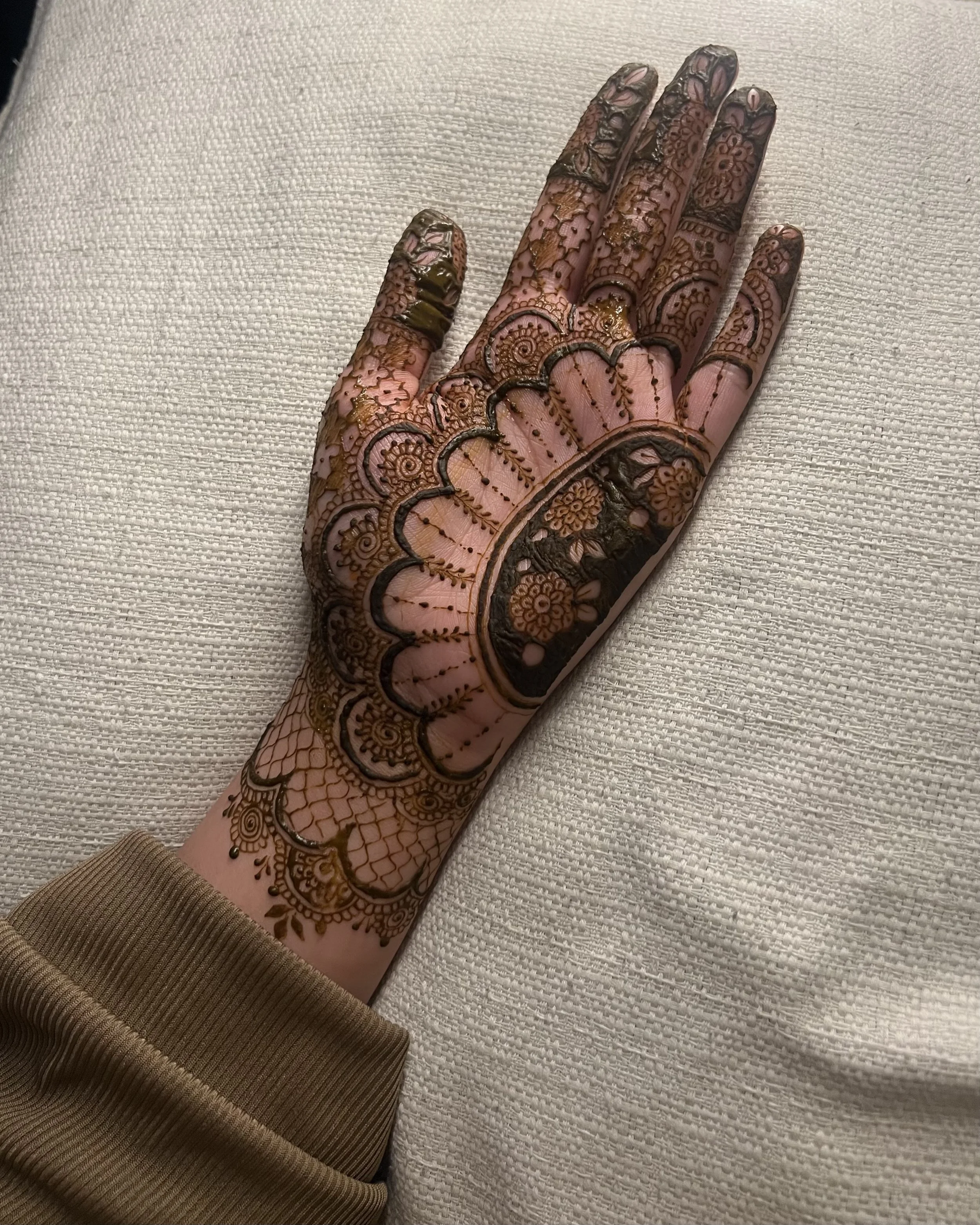 Hand decorated with intricate henna designs resting on a textured white fabric surface, wearing a brown long-sleeve garment.