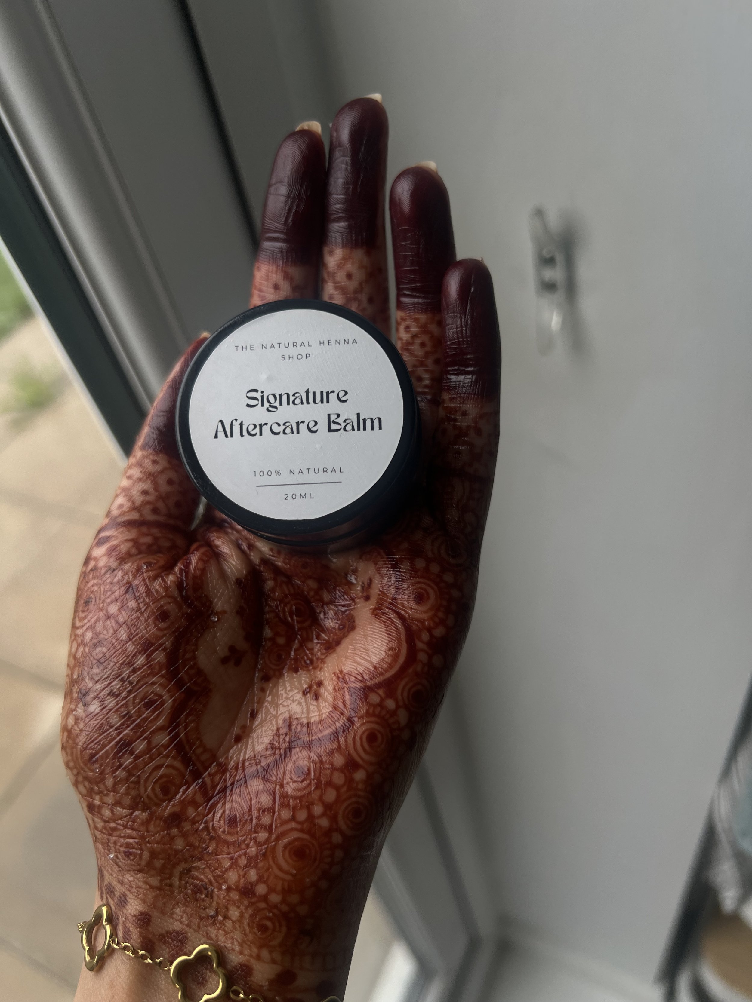 A person's hand with a dark henna / mehndi stain, holding a small jar of 'Signature Henna Aftercare Balm' by The Natural Henna Shop near a window.