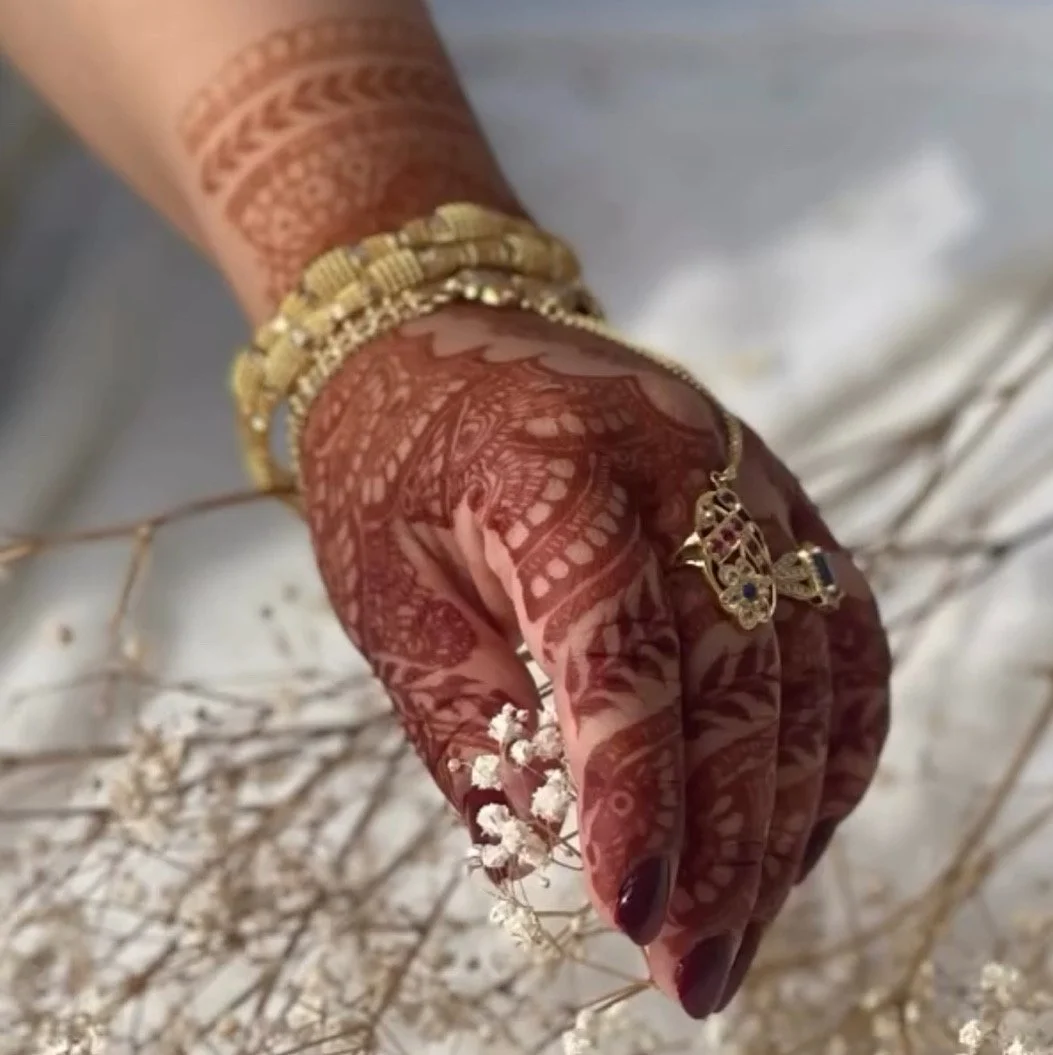 Close-up of a woman's hand adorned with multiple gold rings, a gold bracelet, and intricate henna designs , holding dried white flowers. Dark henna stain using natural henna from the natural henna shop