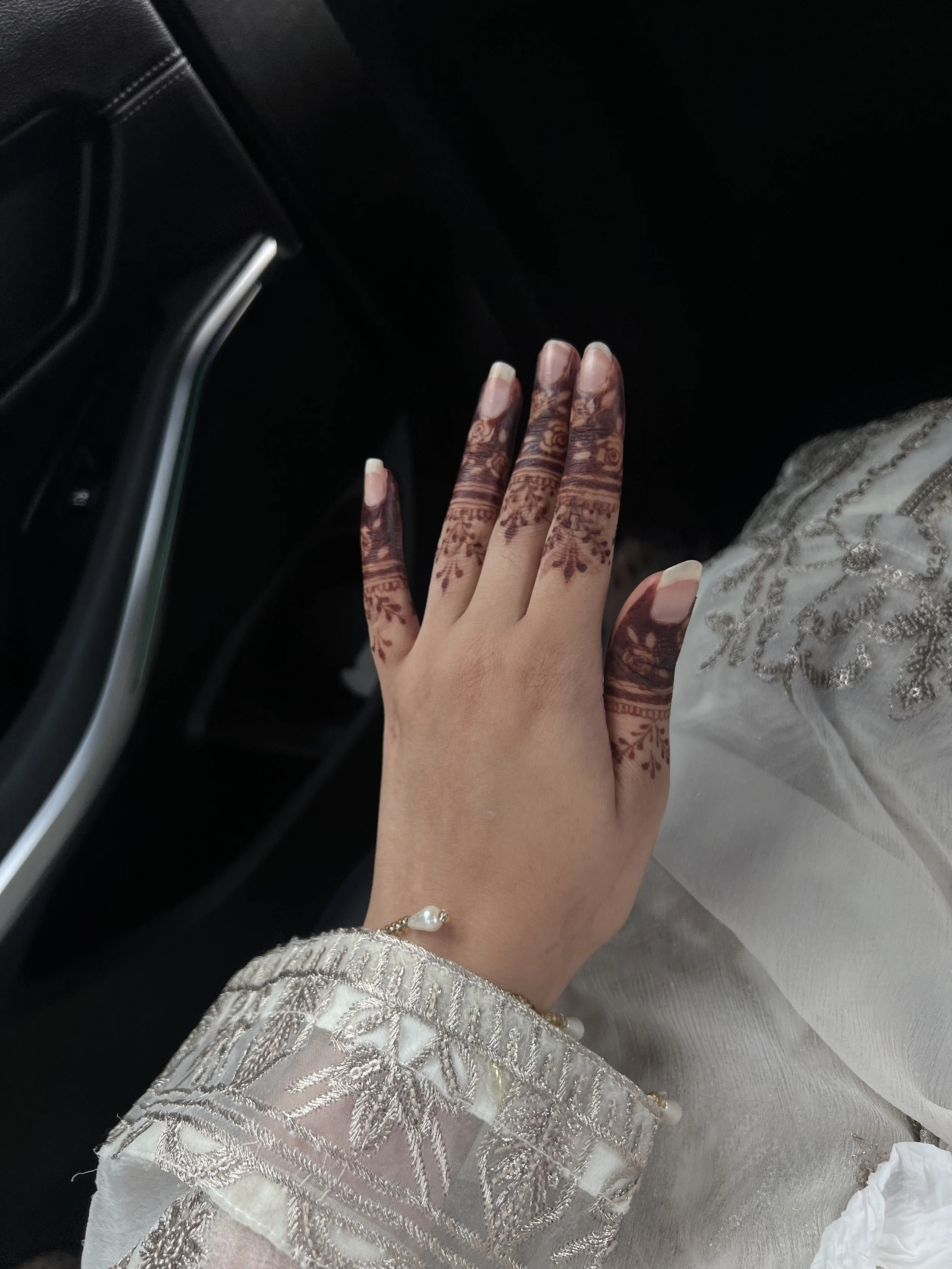 A hand with henna designs resting on a car seat, wearing a gold bracelet with a pearl, and dressed in a cream-colored or white embroidered traditional outfit.
