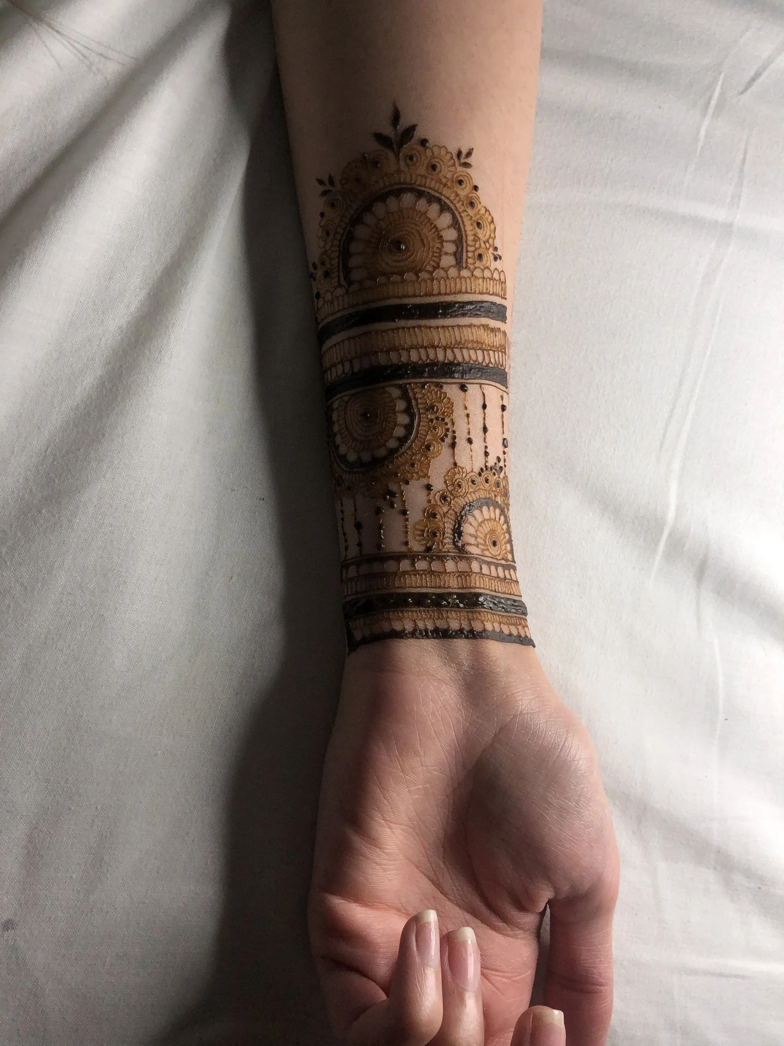 A person's arm and hand with traditional henna tattoo designs on the forearm, featuring intricate patterns and floral motifs, resting on a white surface.