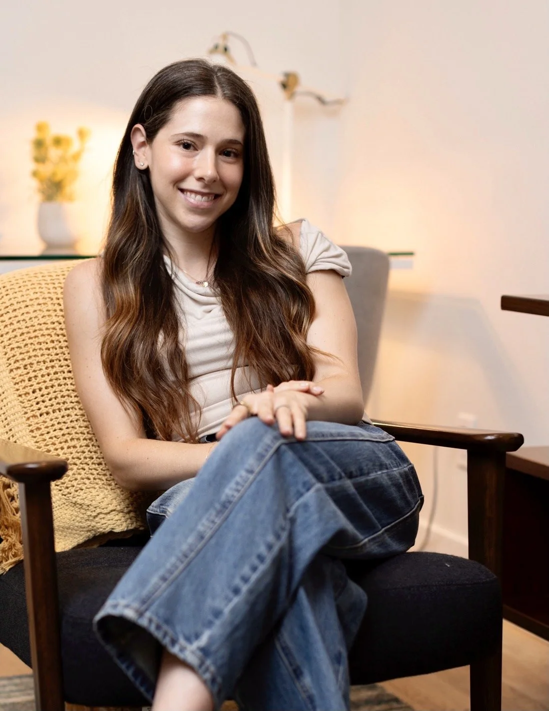 A young woman with long brown hair sitting on a black and wooden chair, smiling at the camera, in a cozy, modern room with warm lighting, a yellow throw pillow, and a potted plant in the background.