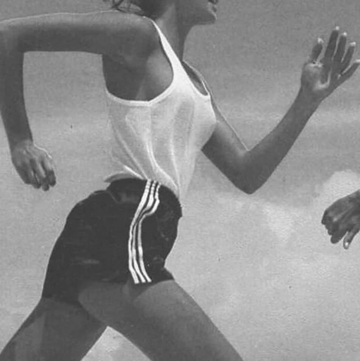 A woman running outdoors wearing a white tank top and black athletic shorts with white stripes.