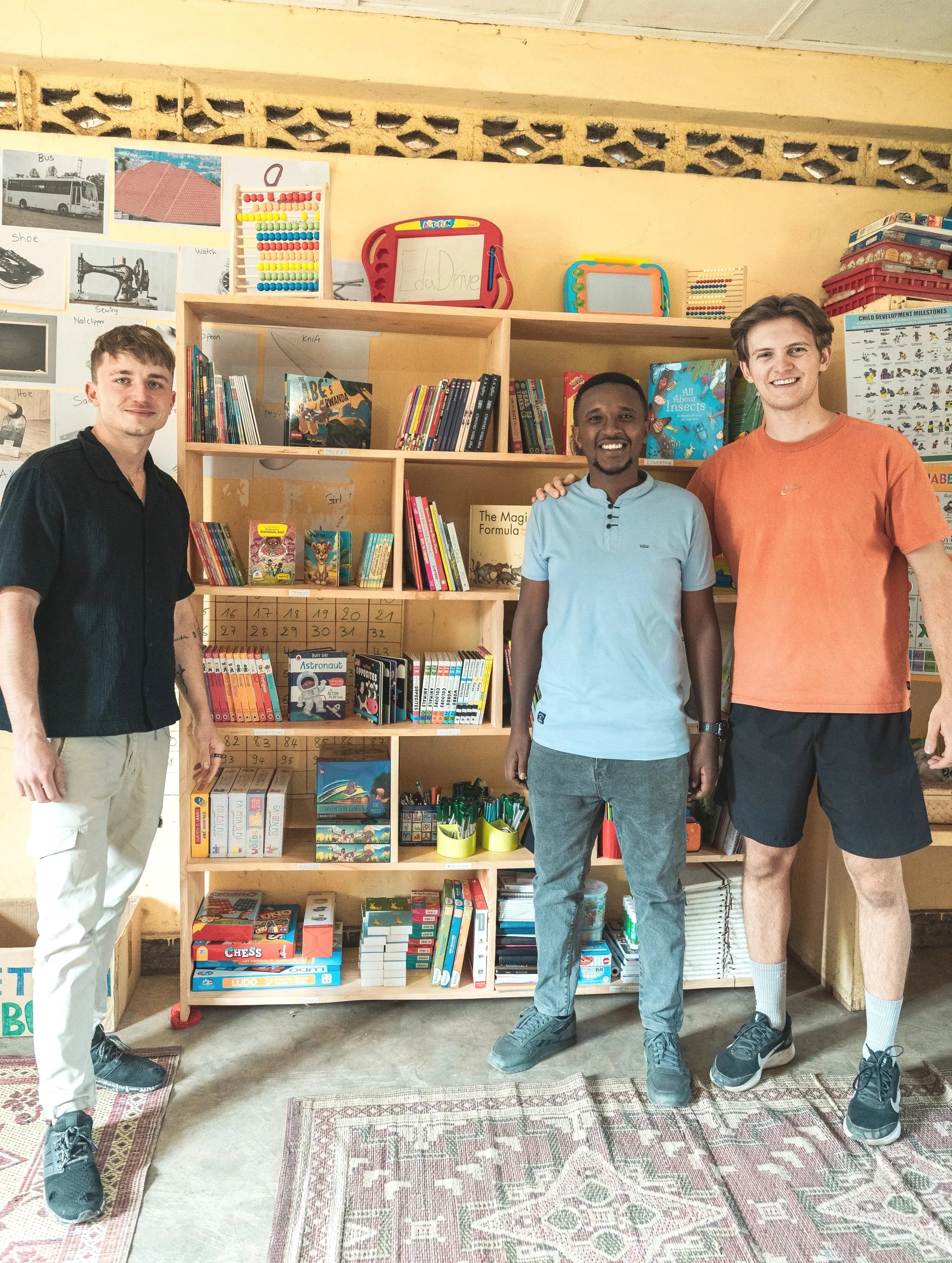 Three men smiling in a library or classroom, standing in front of a wooden bookshelf filled with children's books and educational materials.