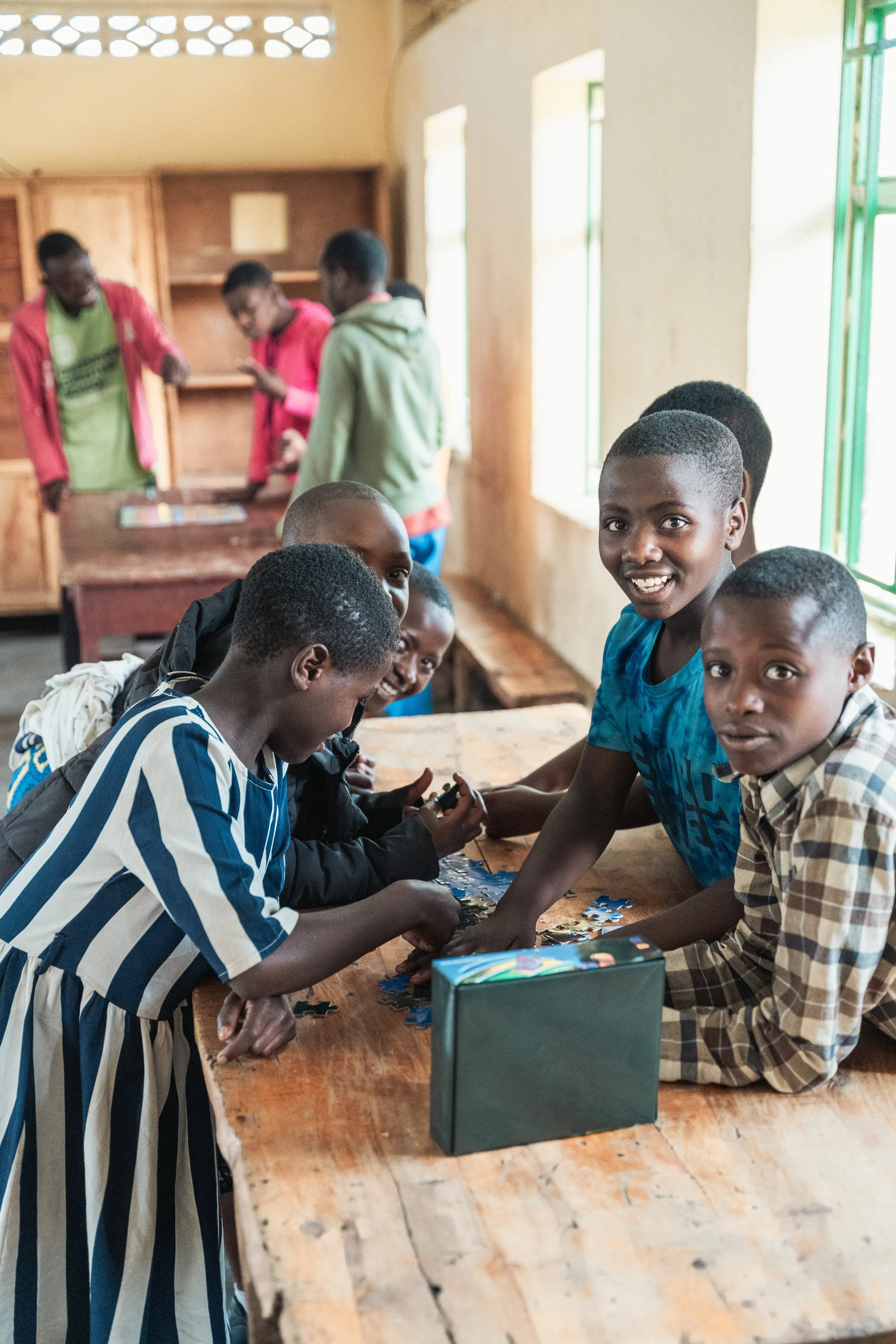 A group of school-aged children working together on a puzzle in a classroom with wooden walls and windows.