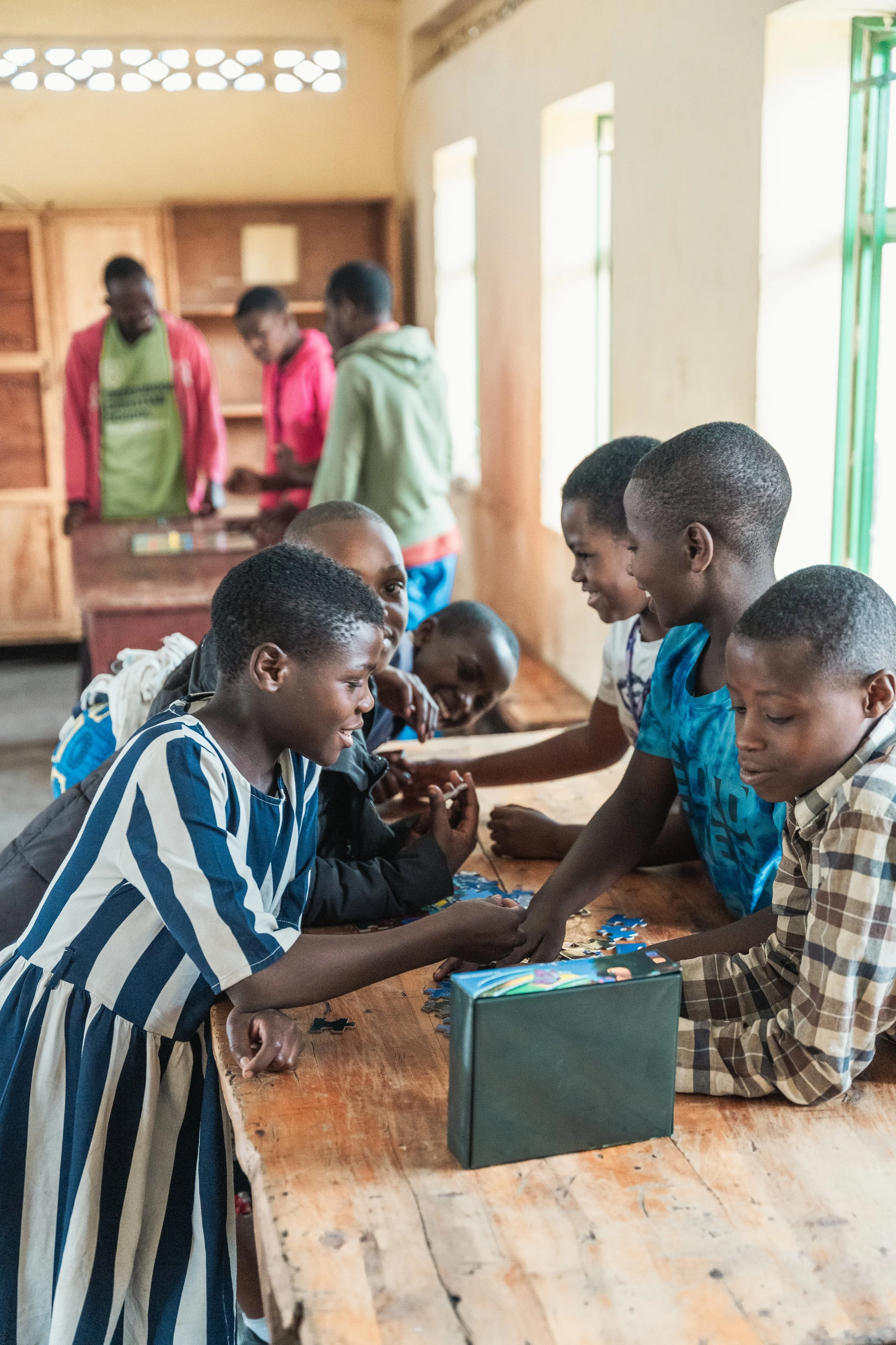 Group of children and teenagers playing with a puzzle on a wooden table inside a classroom.