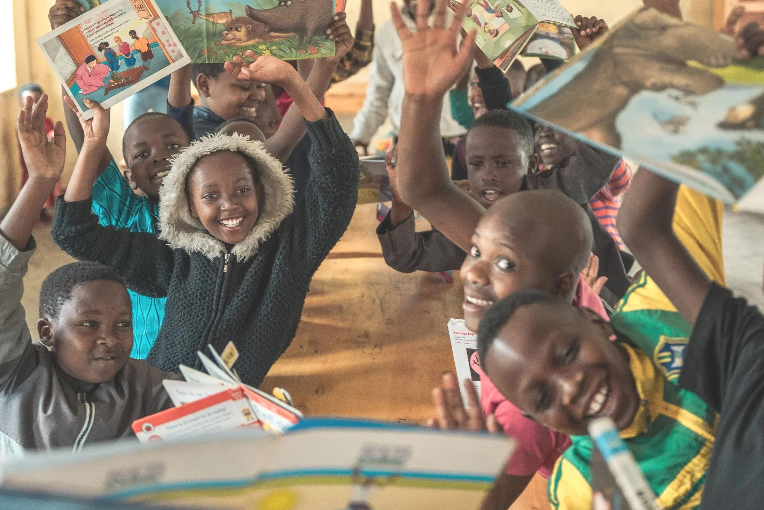 A group of children sitting at a wooden table, happily raising their books and smiling during a reading activity in a classroom.