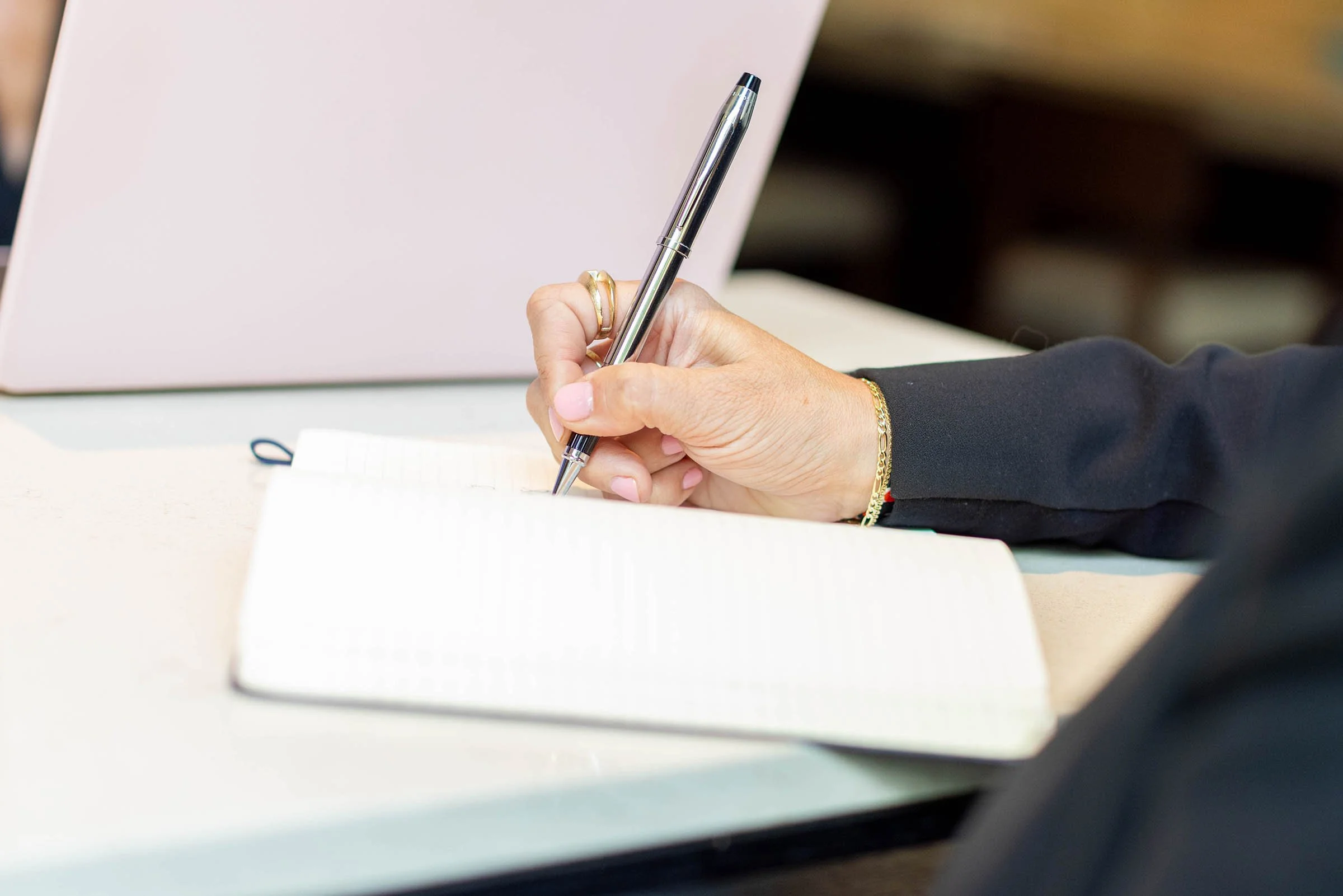 A person writing on a notepad with a silver pen, seated at a table with a pink laptop nearby.