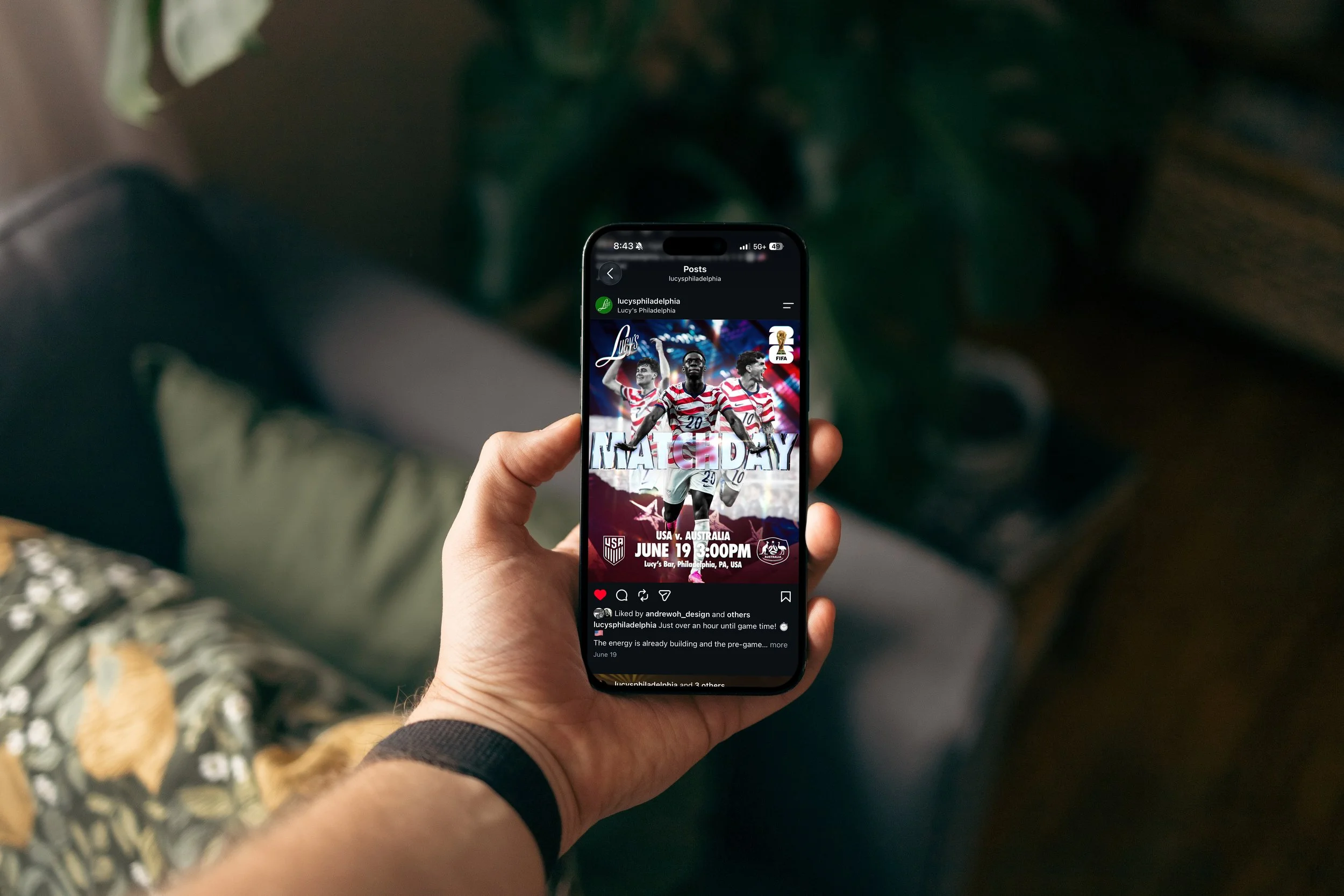 A person holding a smartphone displaying a sports event poster for a soccer match between USA and Australia, scheduled for June 19 at 3:00 PM at Lucy’s Bar in Philadelphia, PA, USA, with a background of blurred indoor setting.