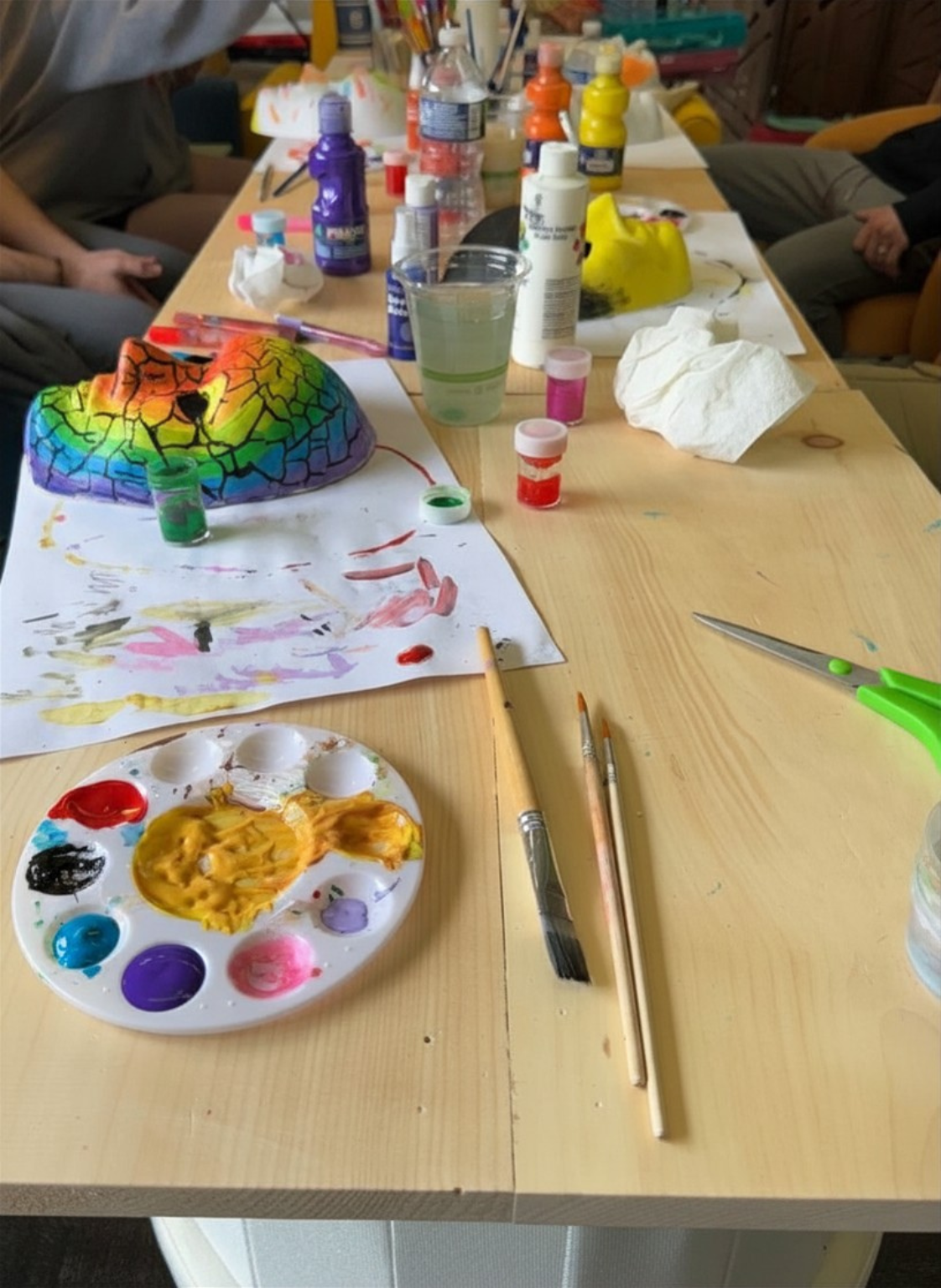 Warm, lived-in expressive arts table during a healing group session. Bright acrylic paints, brushes, scissors, plaster masks, and rainbow crackle art scattered across a communal creative workspace filled with color, process, and human presence.