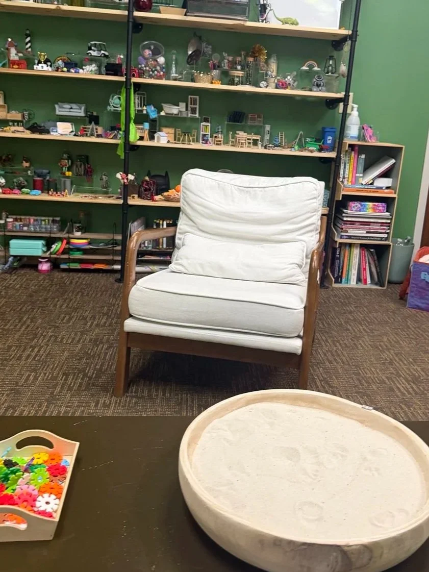Warm expressive arts therapy office with green walls, sand tray, sensory tools, art supplies, and symbolic miniatures creating a playful, grounding, and inviting space.
