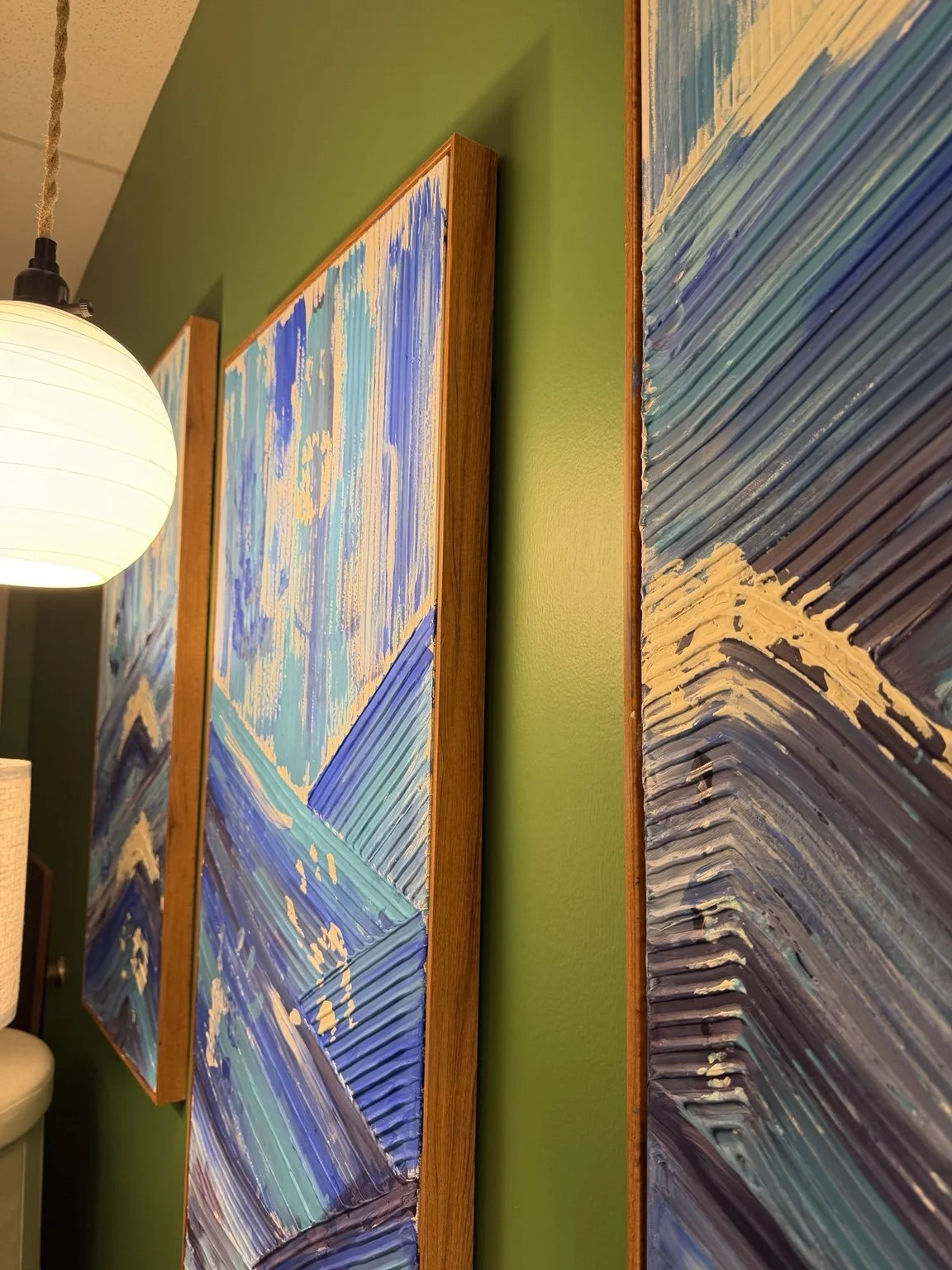 Textured blue triptych painting with layered wave-like brushstrokes in indigo, teal, and white against deep green walls. Warm lighting creates a calming, immersive, ocean-inspired atmosphere.