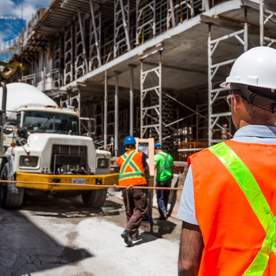 Construction site with workers wearing safety vests and helmets, and a concrete mixer truck in front of partially constructed building.