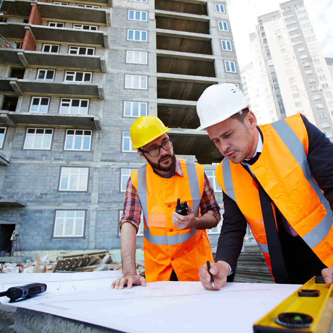 Two construction workers, wearing yellow and white safety helmets and orange safety vests, review blueprints at a building site with a multi-story building in the background.