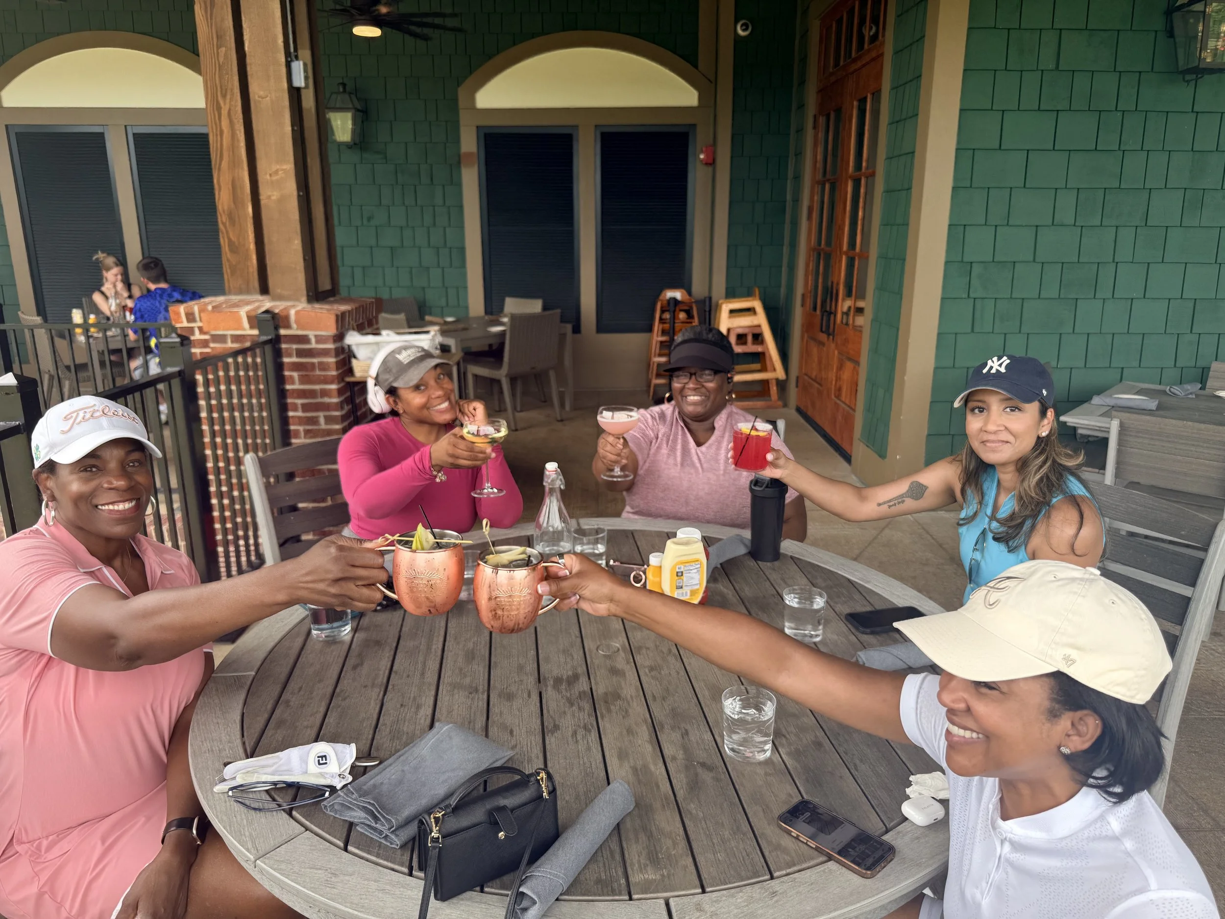 A group of women enjoying drinks at an outdoor restaurant table, smiling and toasting, with one woman wearing headphones, in a casual setting with a brick and green siding background.