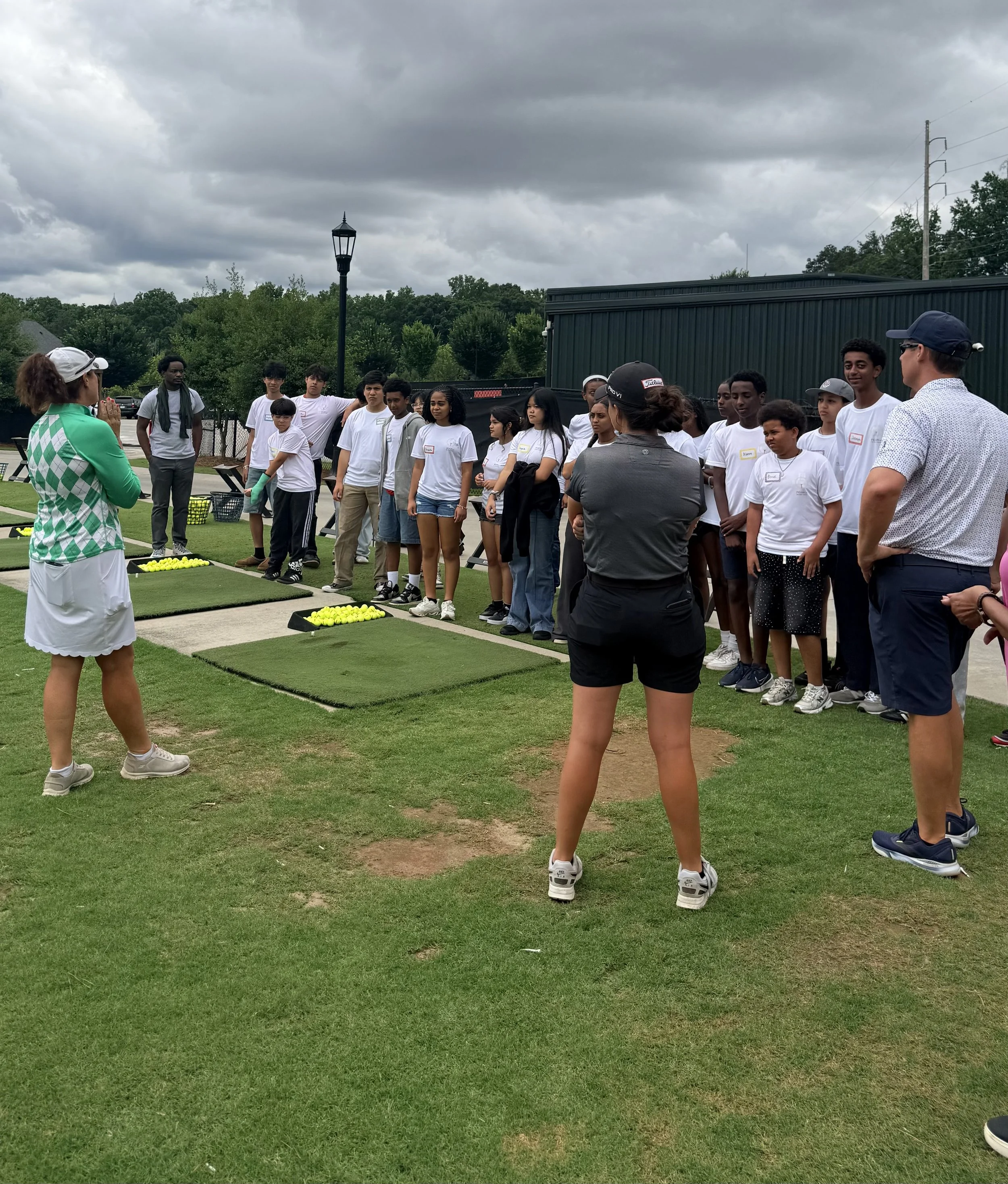 A group of young people standing outdoors on a cloudy day, listening to a woman in a green and white shirt explaining tennis techniques, with tennis balls on a mat in front of her.