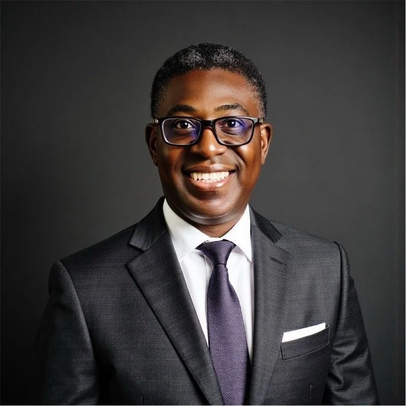 Portrait of a smiling man in a suit with glasses against a dark background.
