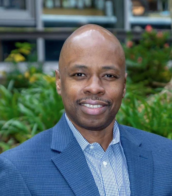 Headshot of a smiling man with a bald head, wearing a blue checked dress shirt and a blue blazer, standing outdoors with green plants and a building in the background.