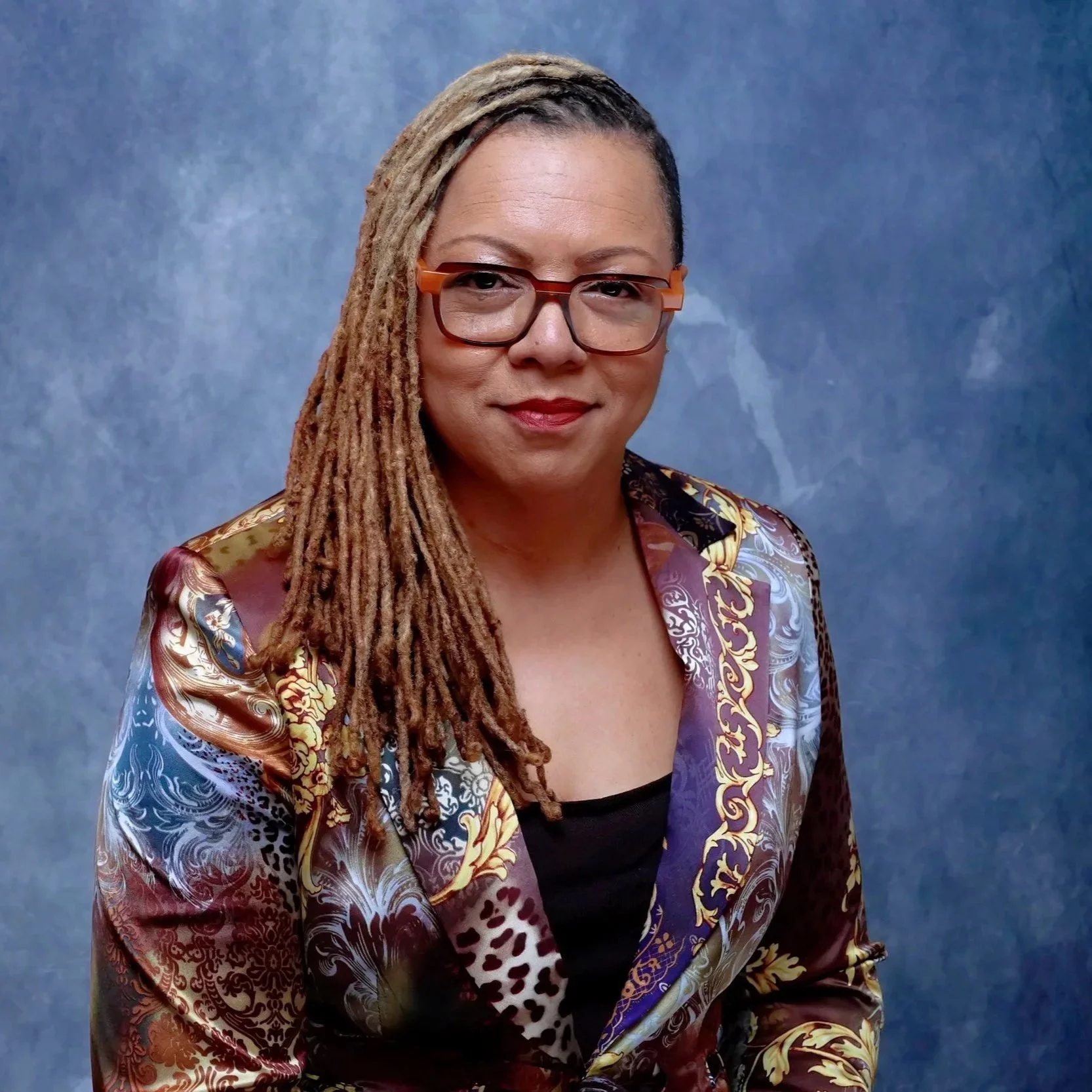 A woman with long dreadlocks, red glasses, and a colorful blazer standing against a blue textured background.
