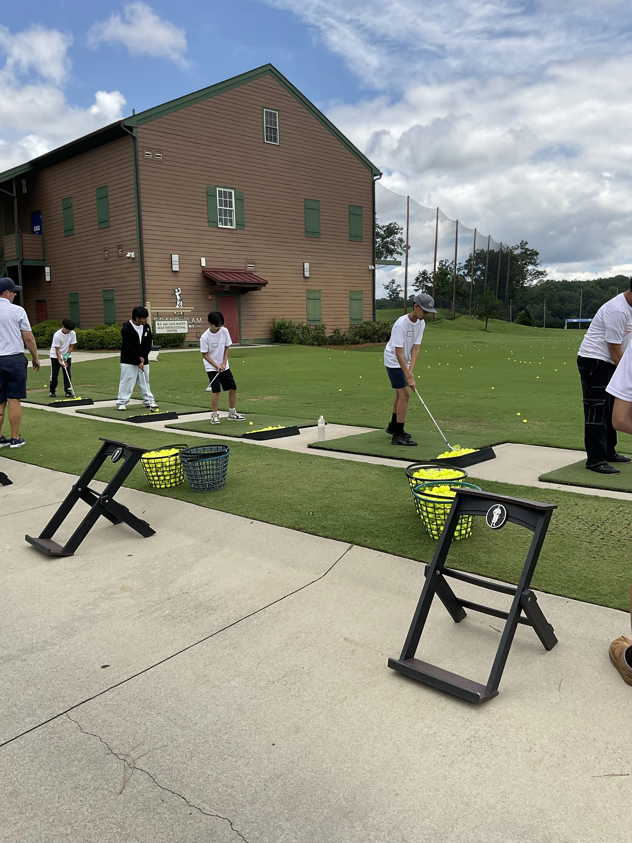 People practicing golf on a driving range with yellow golf balls, baskets, and mats, next to a large brown building with green shutters and a cloudy sky.