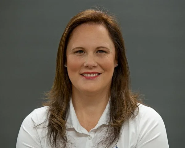 A woman with brown hair smiling, wearing a white shirt, in front of a plain gray background.