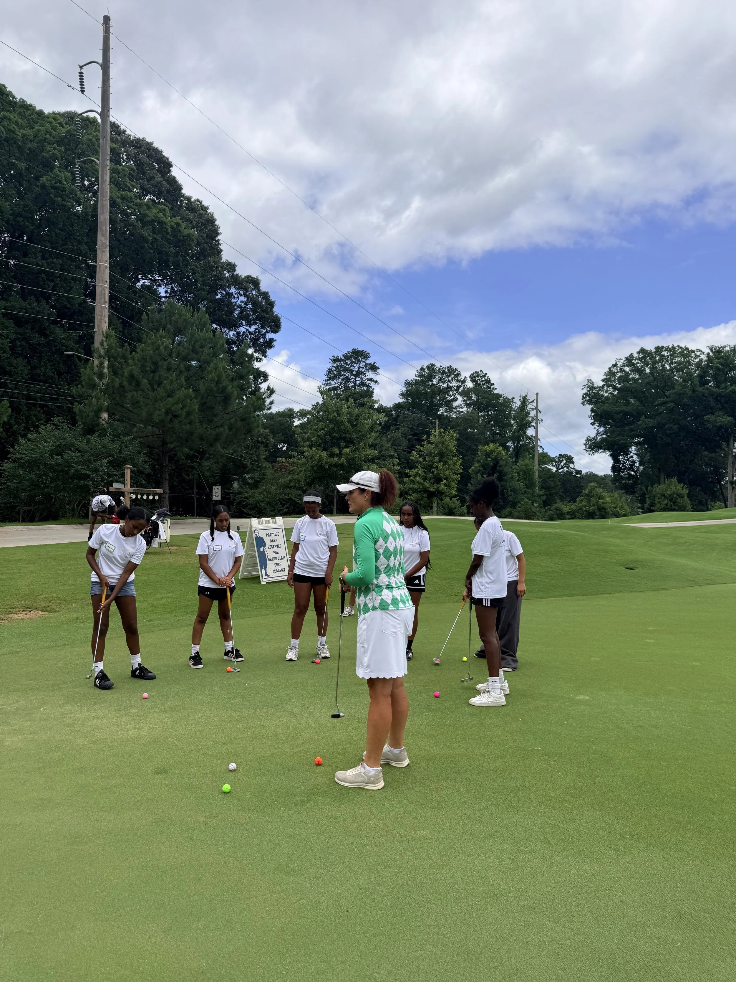 A group of young girls on a golf course learning to putt, with a woman instructor in a green and white shirt guiding them. There are colorful golf balls on the grass and a sign in the background.