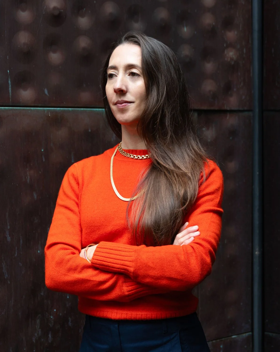 A woman with long, wavy brown hair, wearing an orange sweater and gold jewelry, stands with arms crossed against a dark, textured background.