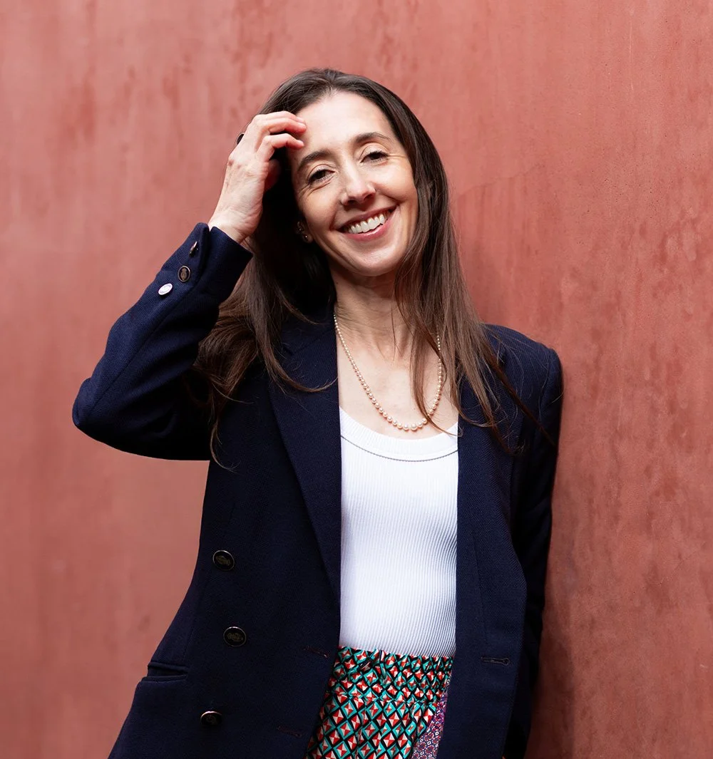A woman with long brown hair smiling and touching her hair, wearing a navy blue blazer, a white top, is standing against a textured pink wall.