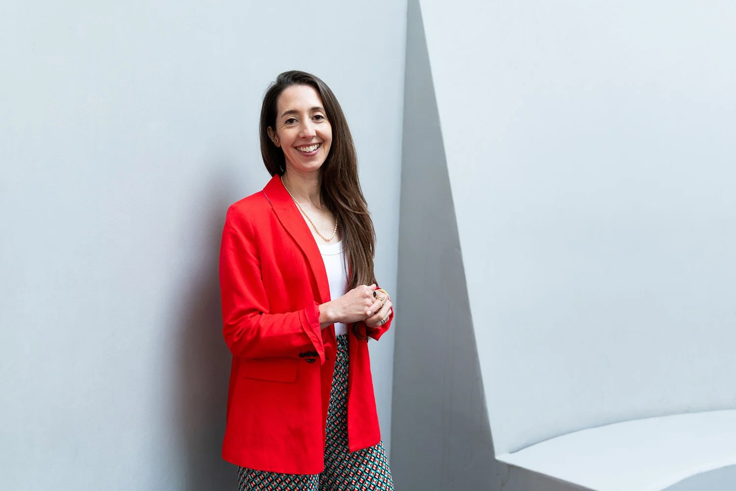A smiling woman with long brown hair wearing a red blazer, white top, and patterned pants, standing against a light-colored wall in an indoor setting.