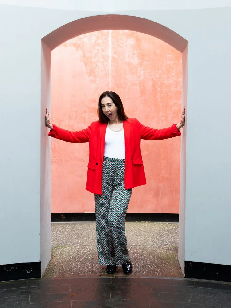 Woman in red blazer and patterned pants standing in a doorway with a pink and gray background.