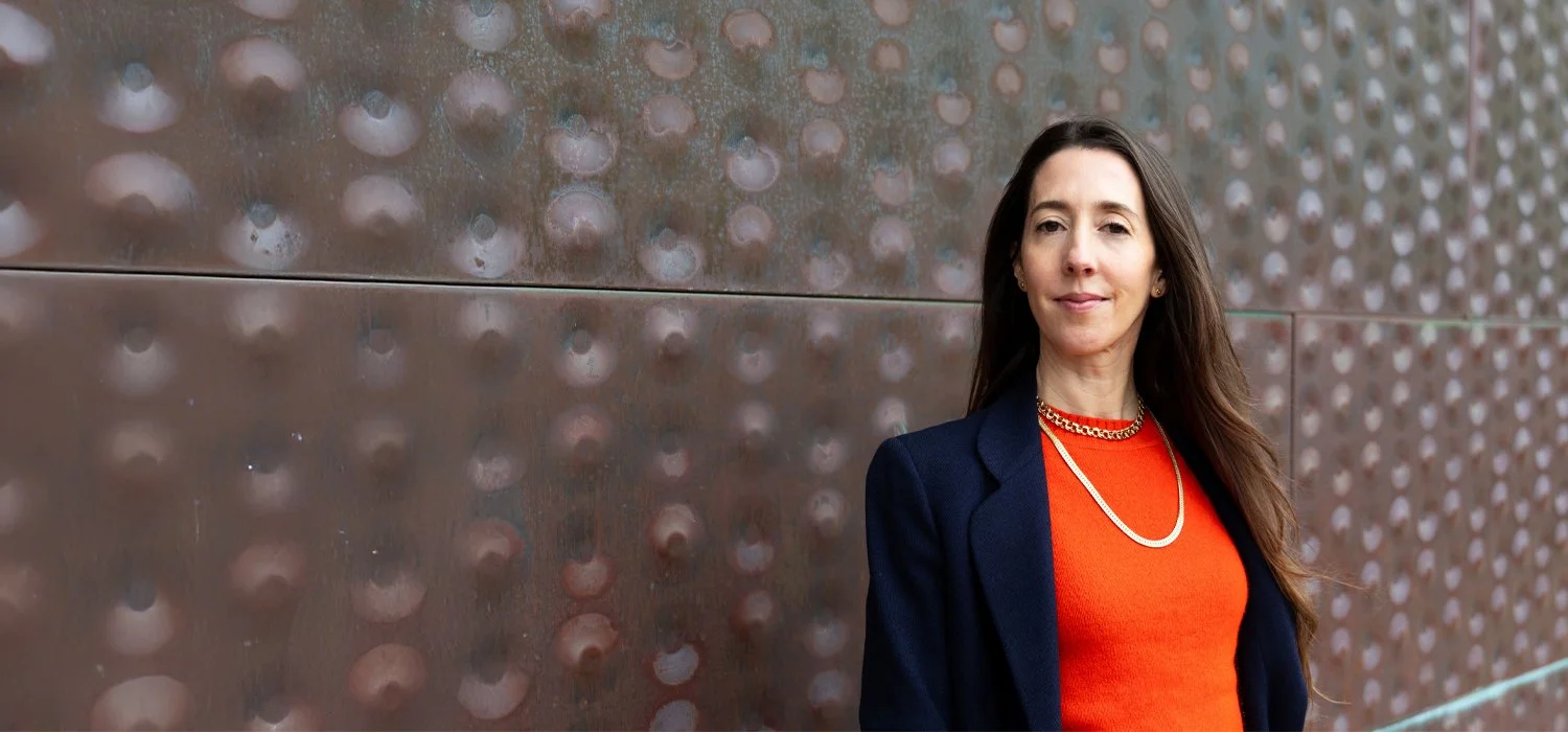 Woman with long brown hair wearing a red top, navy blazer, and gold jewelry standing outdoors against a textured metallic wall.