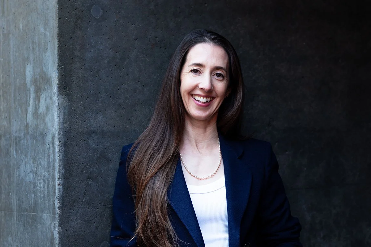 A woman with long brown hair, wearing a navy blazer and a pearl necklace, smiling and standing against a dark and light gray concrete wall background.